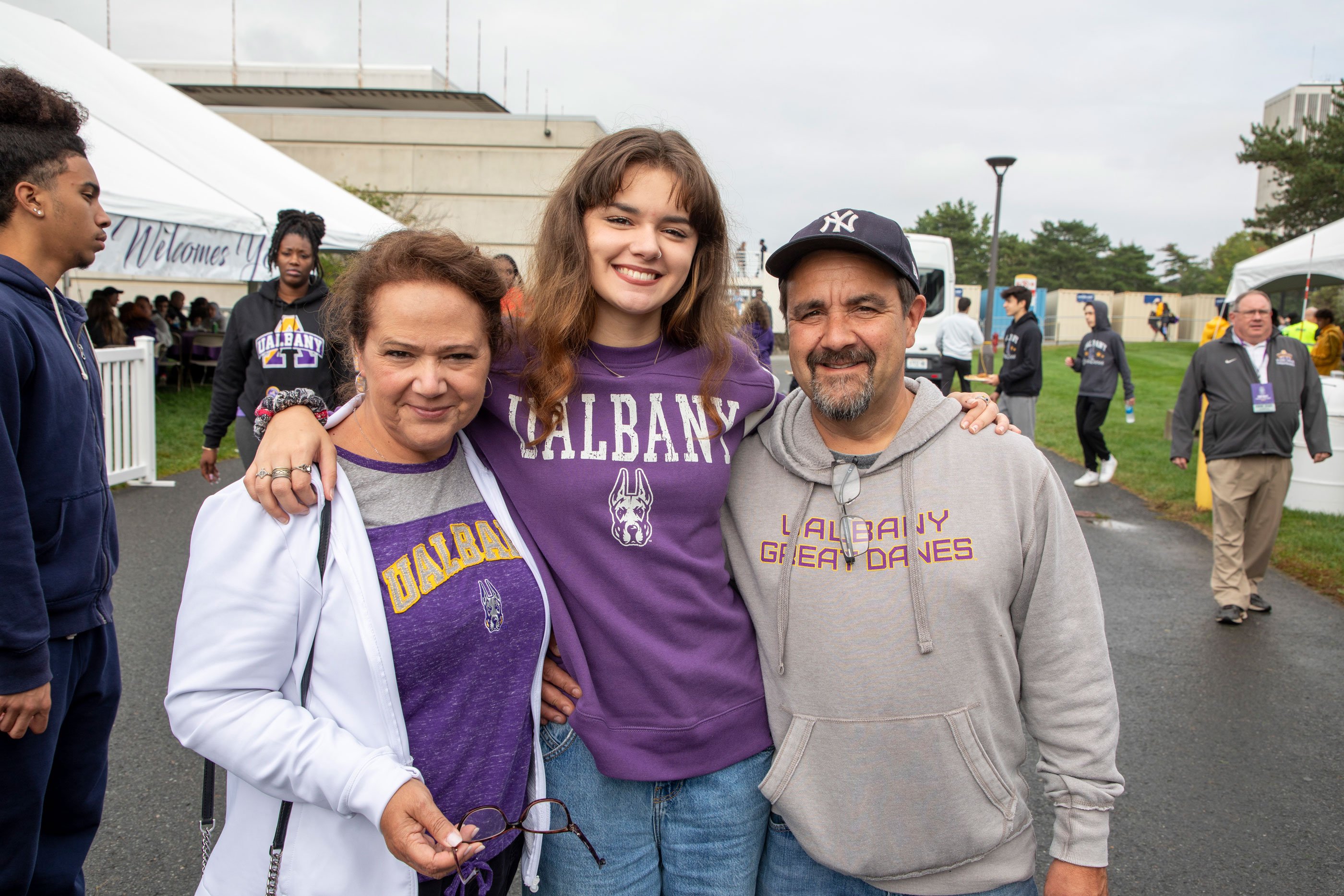 A family of three wearing UAlbany shirts smiles as they pose for a photo.