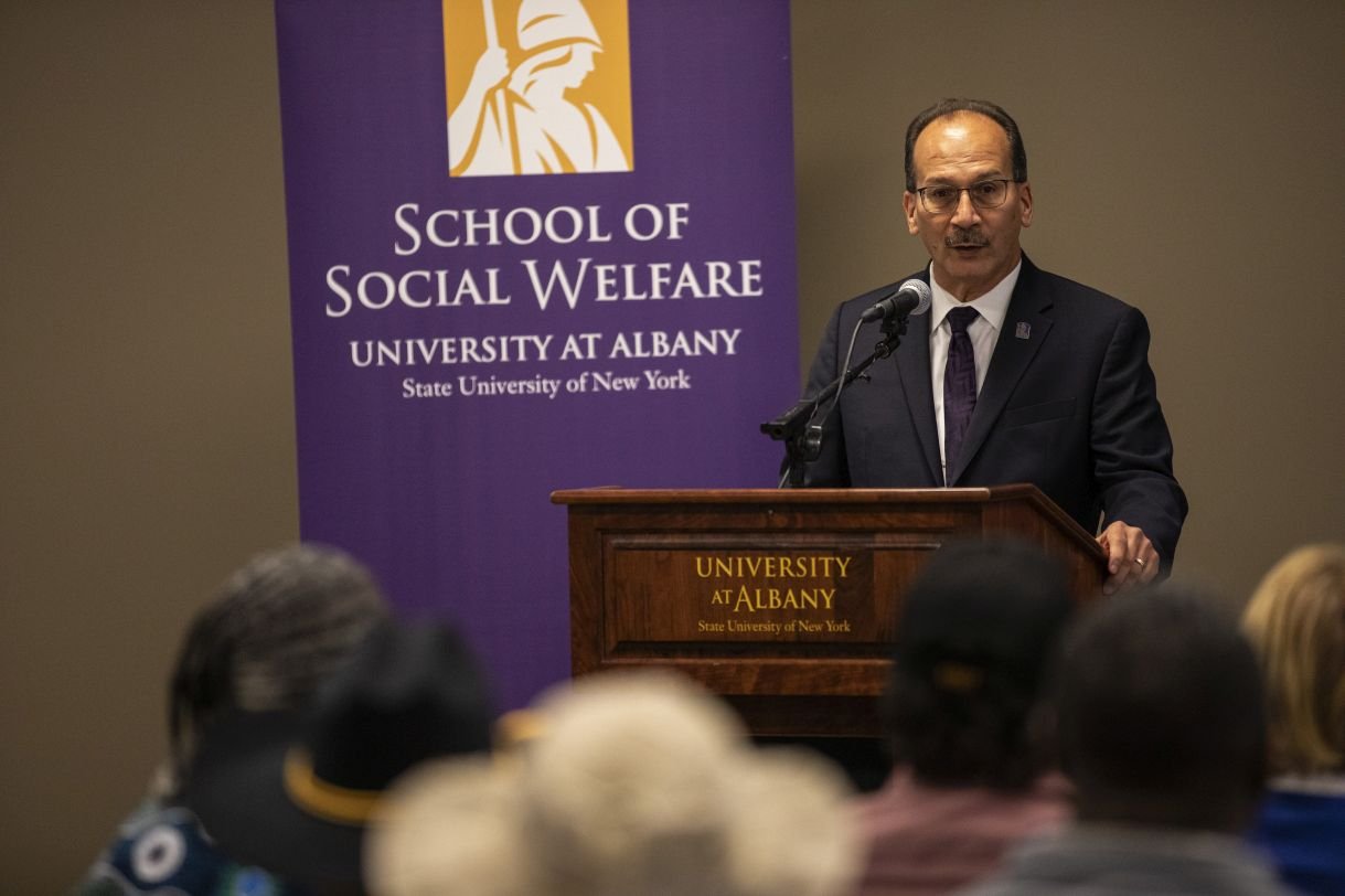 President Havidán Rodríguez addresses the audience, standing in front of a purple banner which says "School of Social Welfare". He is wearing glasses, a dark suit jacket and purple tie.