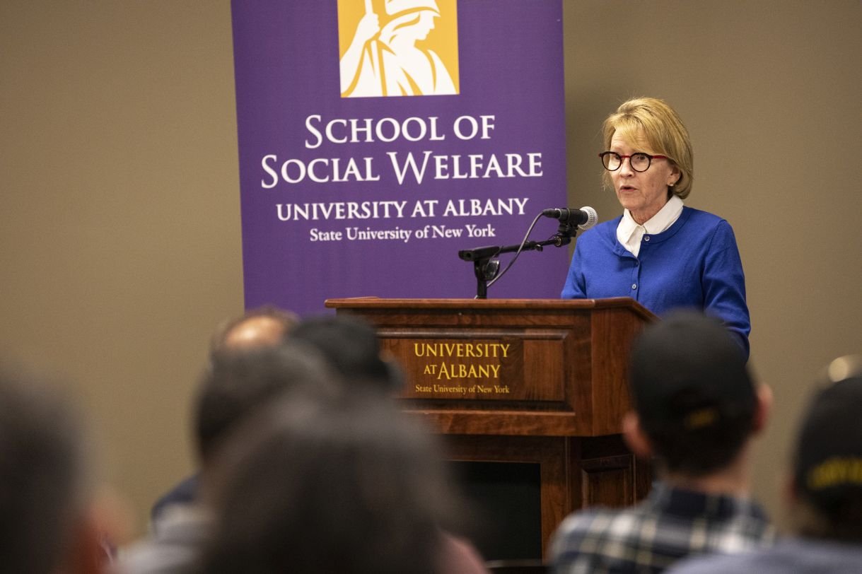  Aileen Gunther delivers remarks at a podium that says "University at Albany", in front of a purple banner which says "School of Social Welfare". She is wearing a bright blue cardigan over a white collared shirt with dark rimmed glasses.