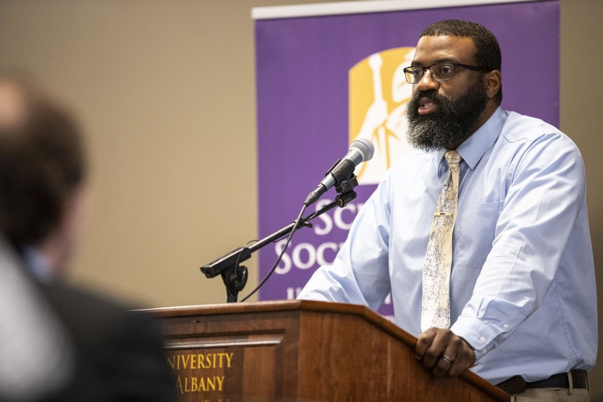 Gavin Walters delivers remarks at a podium that says "University at Albany", in front of a purple banner which says "School of Social Welfare". He is wearing black rimmed glasses, a light blue button up shirt and a pale yellow tie. 