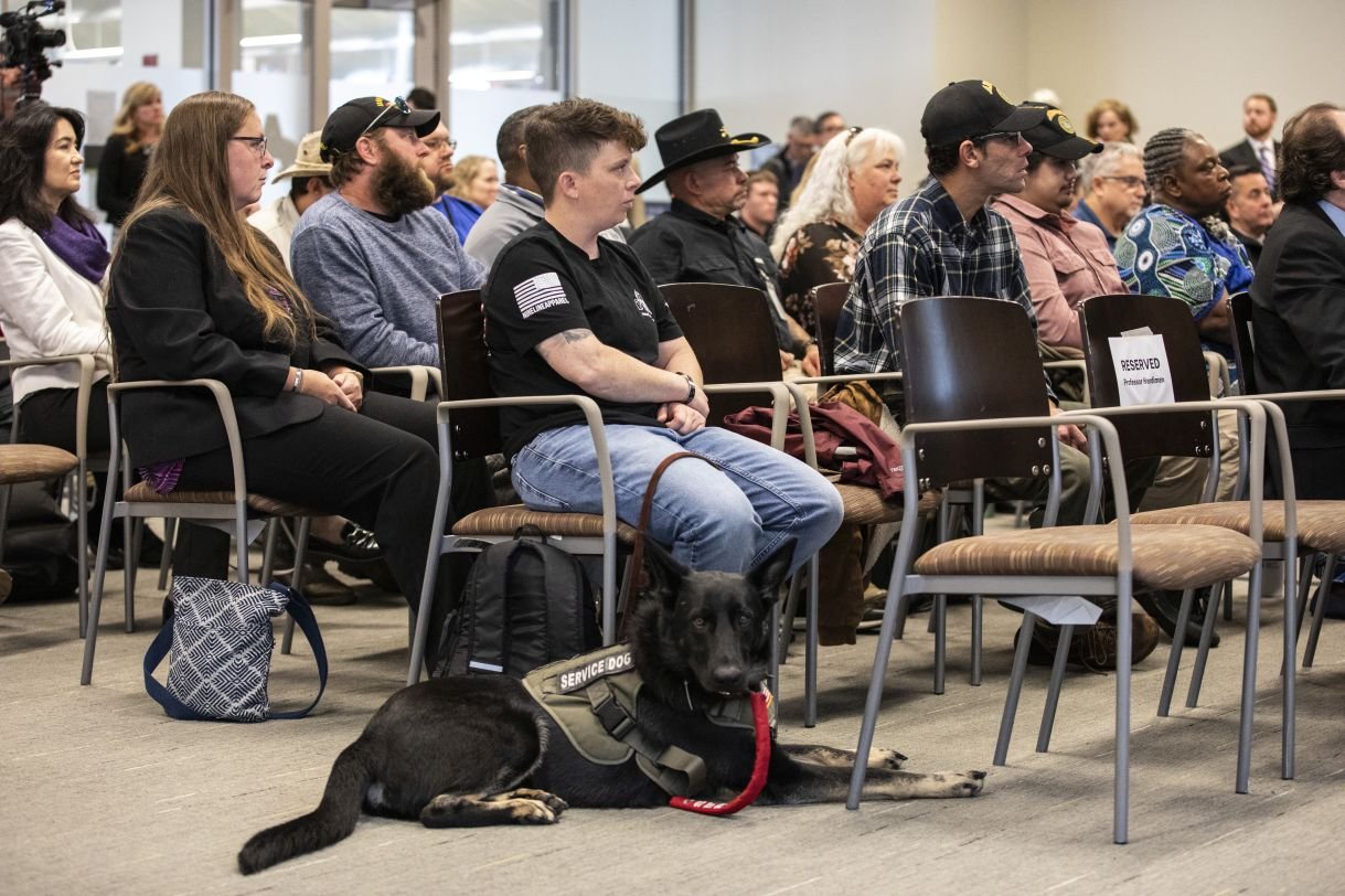 Dwyer program participants sit in chairs arranged in rows at the press conference. The image focuses on a seated veteran with short dark hair, black T-shirt and jeans, who is accompanied by a black service dog wearing a khaki vest and leash. 