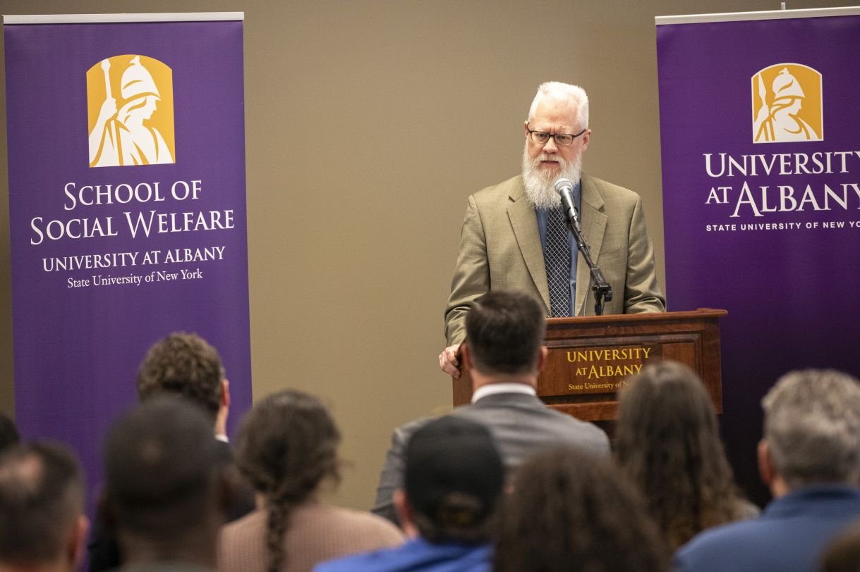 Eric Hardiman delivers remarks at a podium that says "University at Albany", in front of a purple banner which says "School of Social Welfare" and another purple banner which says "University at Albany". He is wearing black rimmed glasses and a tan suit jacket over a gray shirt.