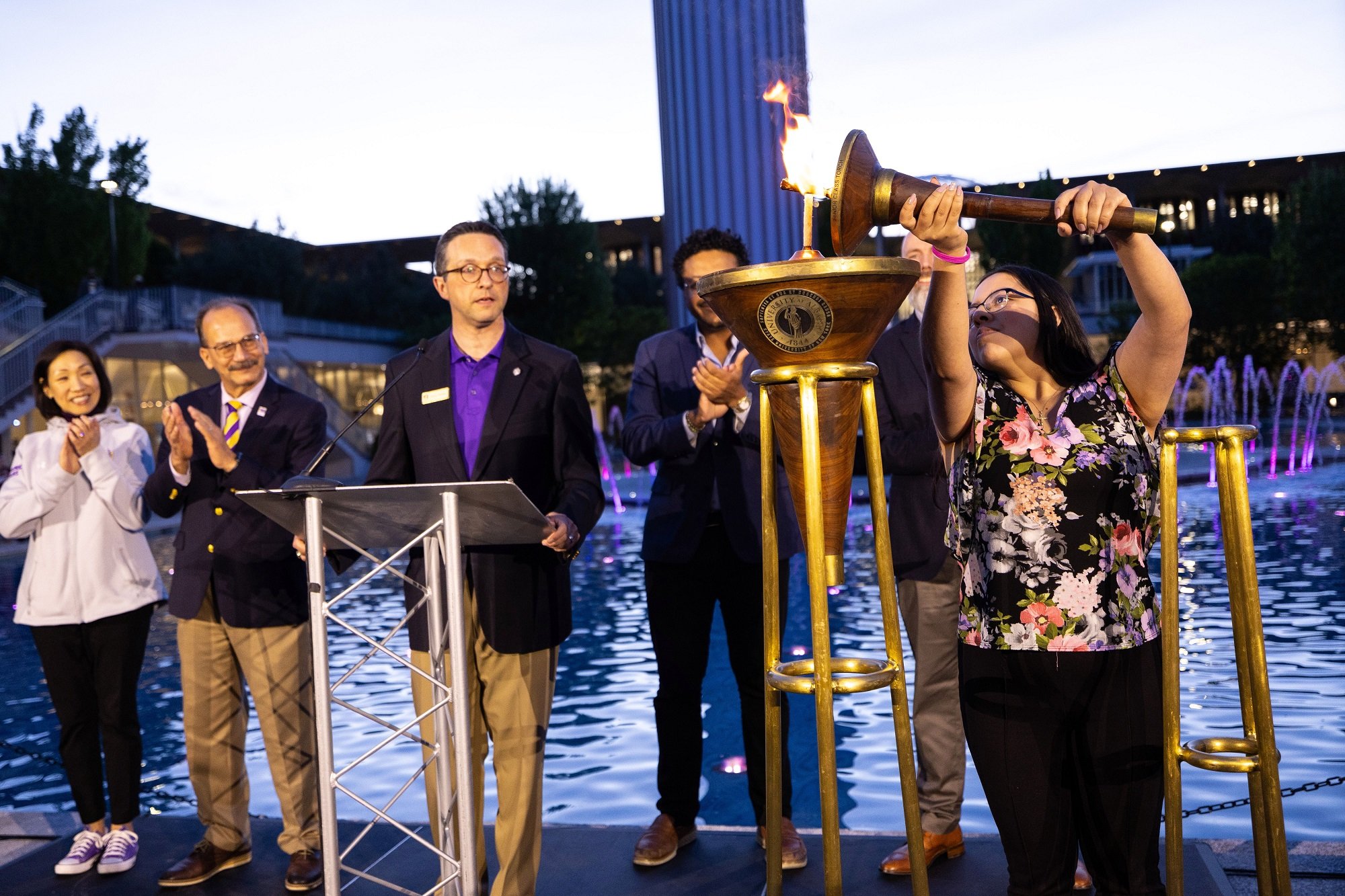 A UAlbany student lights the ceremonial torch at the main fountain alongside UAlbany faculty, staff and alumni.