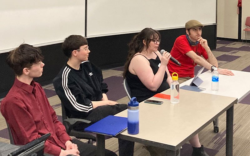 A student sits and holds a microphone alongside three other students while presenting on a panel at UAlbany Showcase