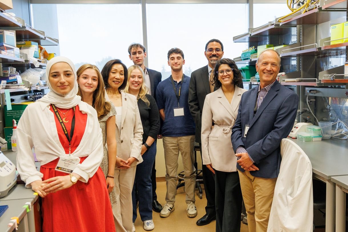 Nine smiling people stand together in a lab, posing for a group portrait. They are flanked by lab benches and shelving stocked with lab supplies. 