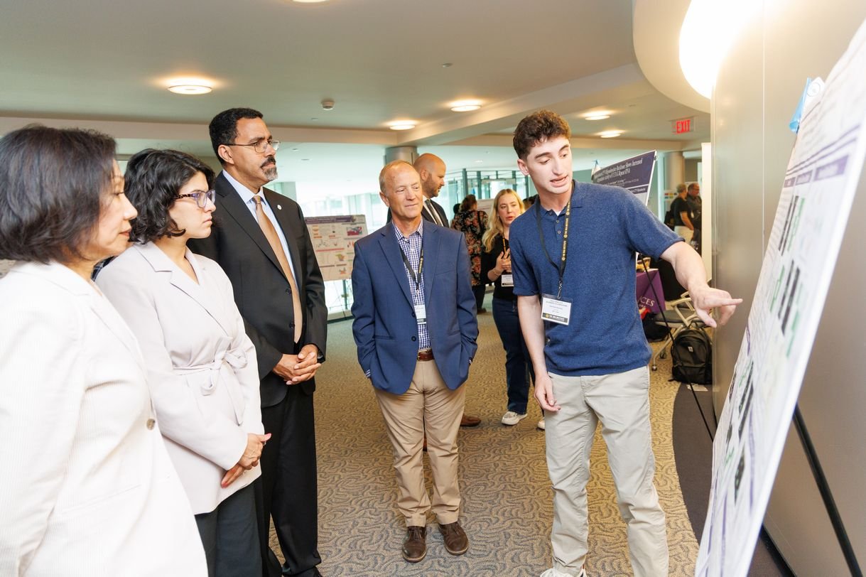 A student explains his research poster to four people. Other people and posters set up on easels are visible in the background.