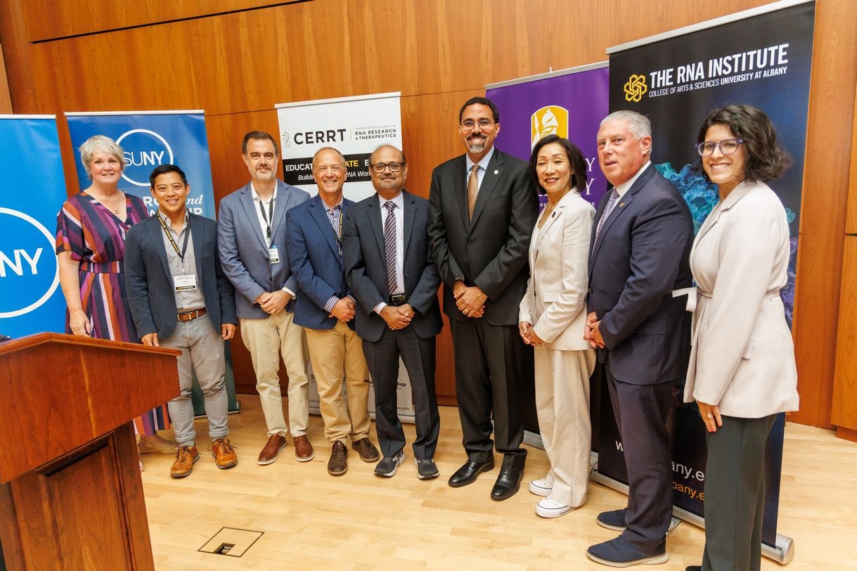 Nine people stand in a row, smiling for a group portrait. They are standing in front of five popup banners that say "SUNY", "CERRT" and "The RNA Institute."