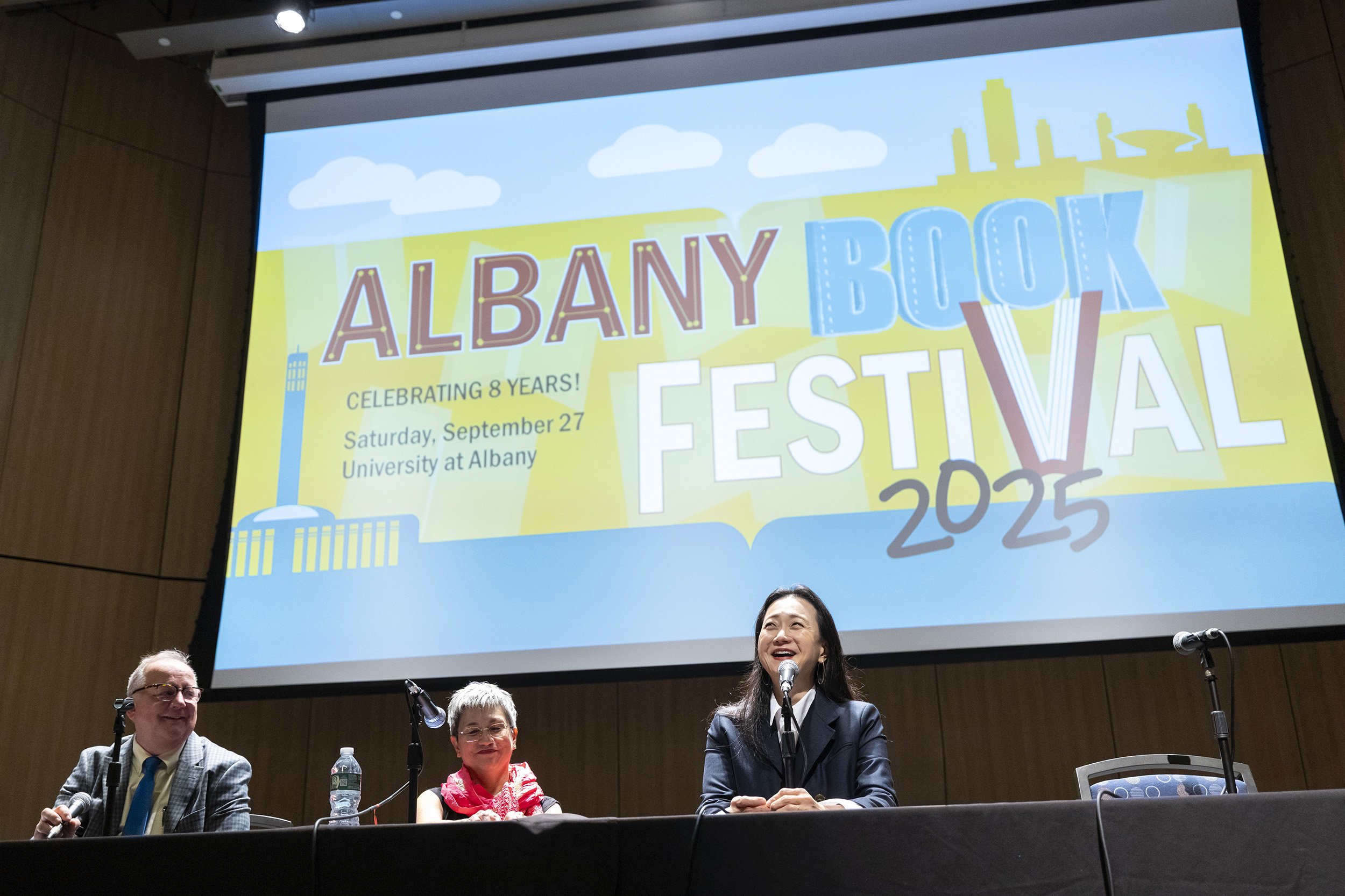 Three people sit in front of a large screen that has the words Albany Book Festival on it indoors in an auditorium.