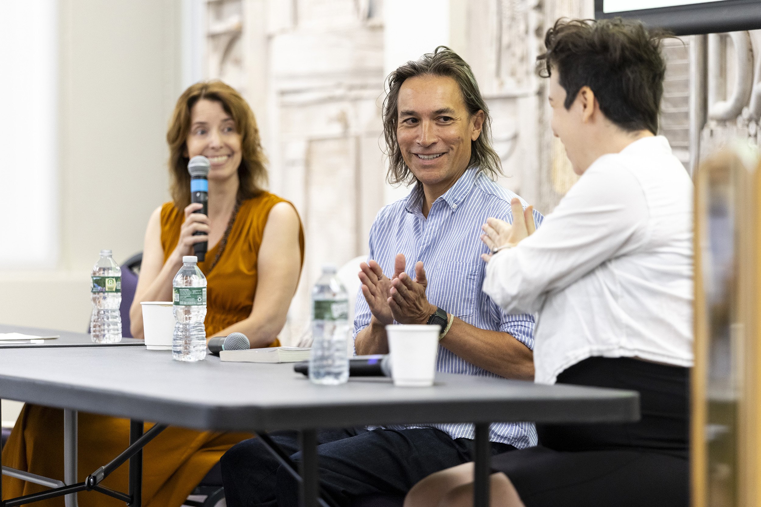 Three people are seated at a table with microphones and water talking with smiles on their faces indoors.