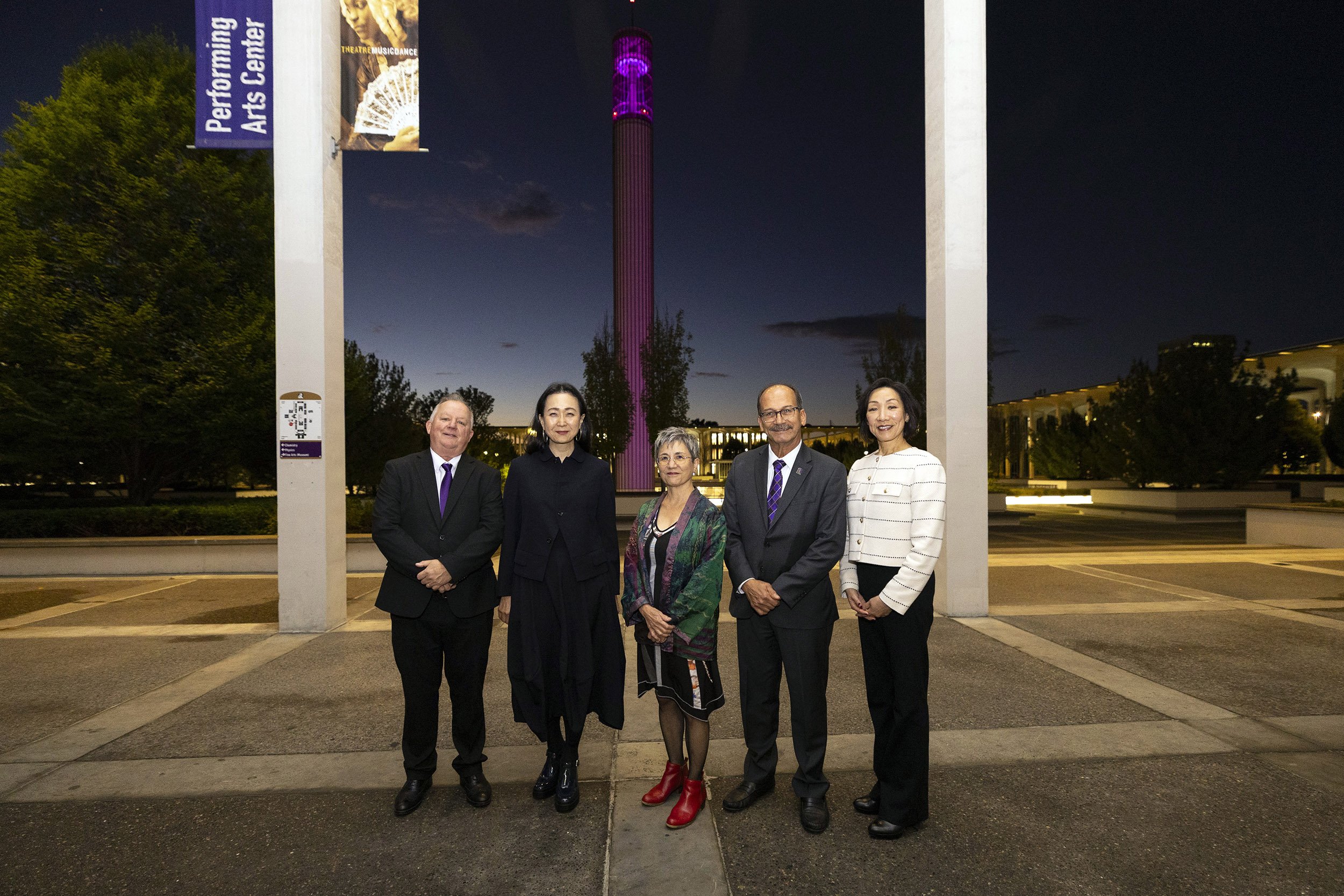 A group of five people stand in a row for a portrait, a man in a suit and purple tie, a woman in a long black coat, a shorter woman in a colorful shawl, a man in a gray suit and purple tie with a mustache and a woman in a white sweater or blazer against a night/dusk skyline with lights in the distance.