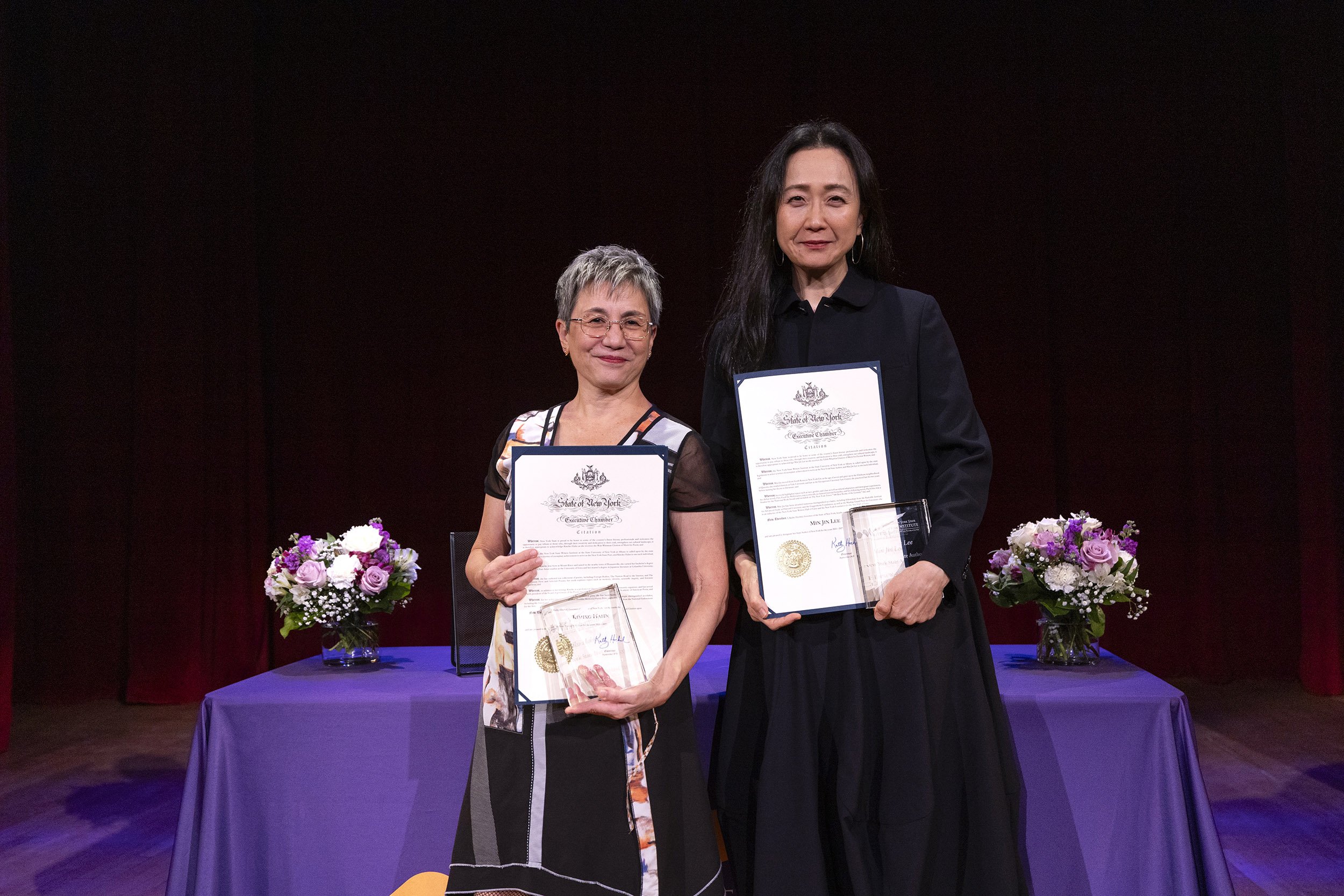 A short woman in a colorful blouse and a tall woman in a dark dress stand with proclamations of State Poet and State Author against a black backdrop and a table with a purple cover.