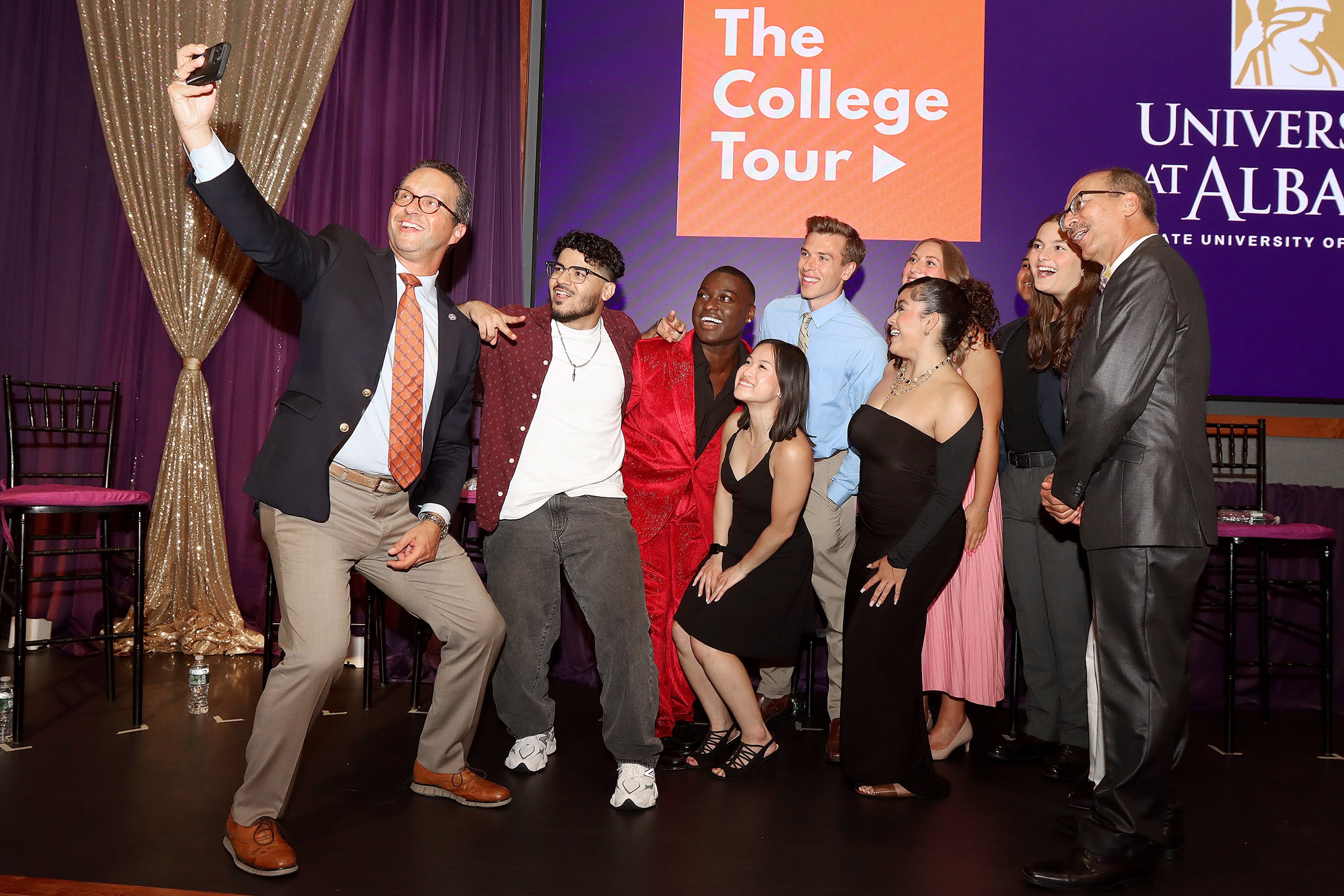 A group of students in colorful outfits pose indoors for a selfie with a man in a blazer and glasses with the words "The College Tour" on the screen behind them.