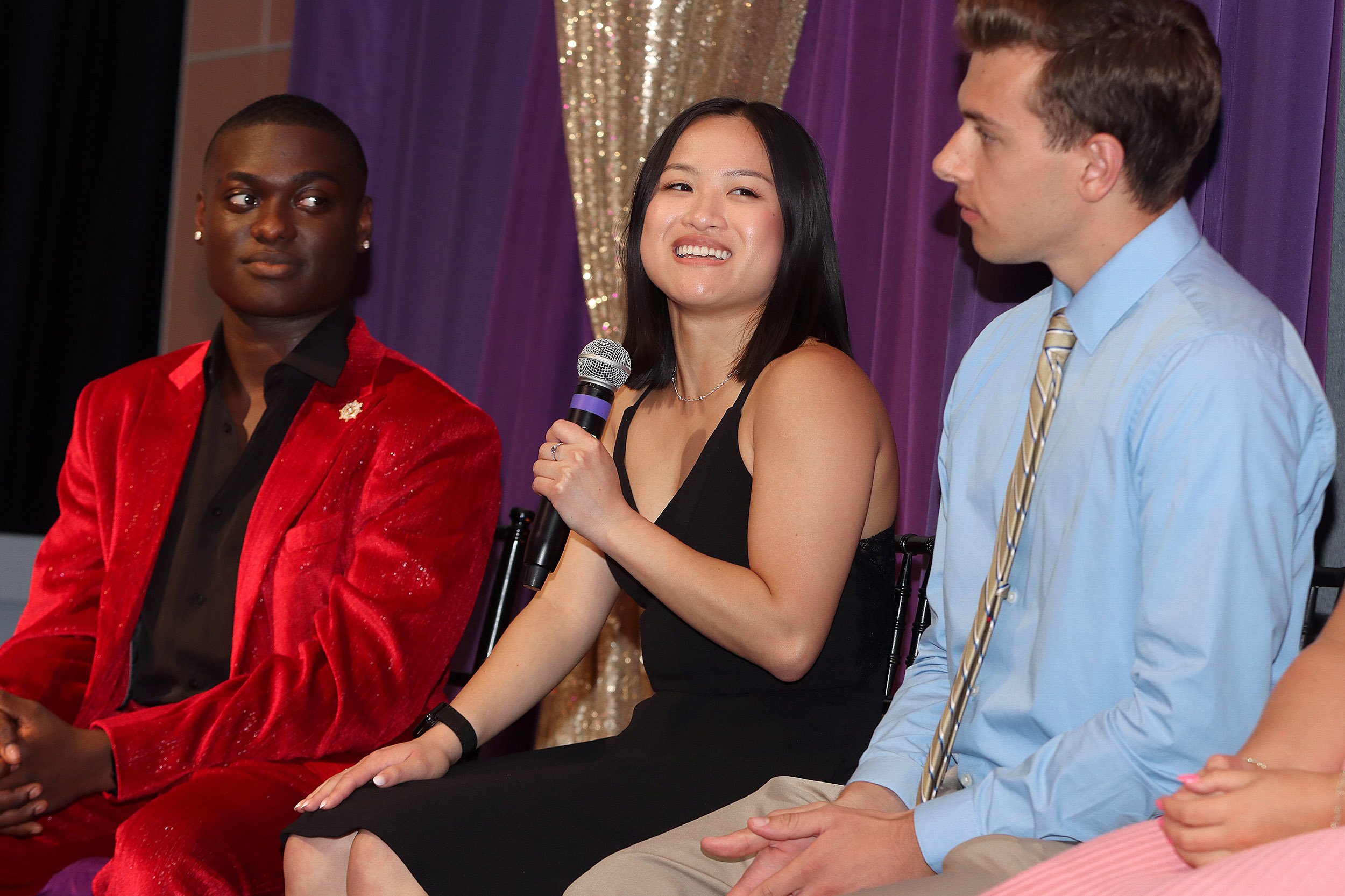A young woman of Asian descent in a black dress is seated holding a microphone in between a young African American man in a red suit and a young white man with brown hair wearing a blue shirt and tie.