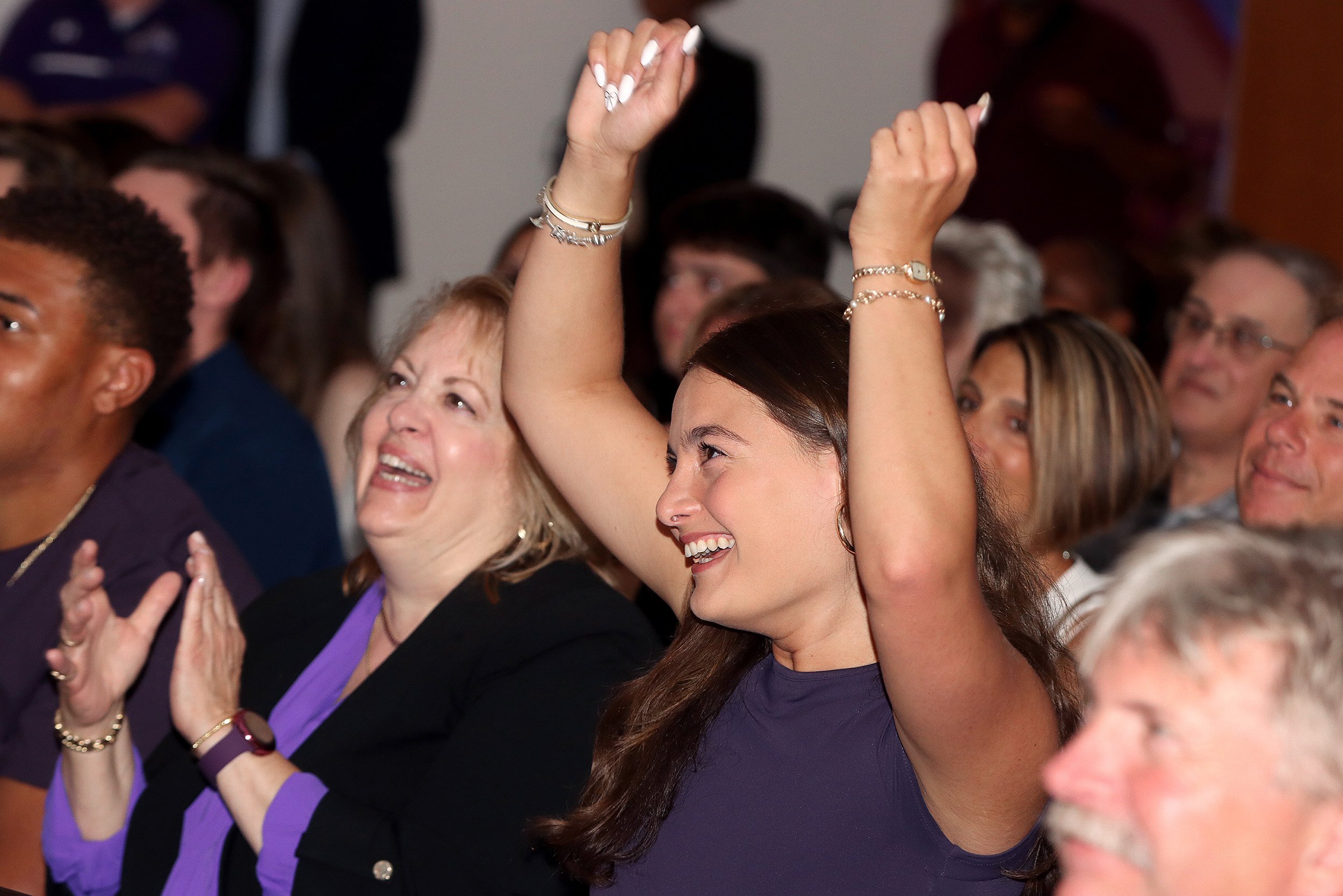 A young woman in a blue top holds her hands up in celebration as a crowd is seated behind her - she looks off screen in front of her indoors.