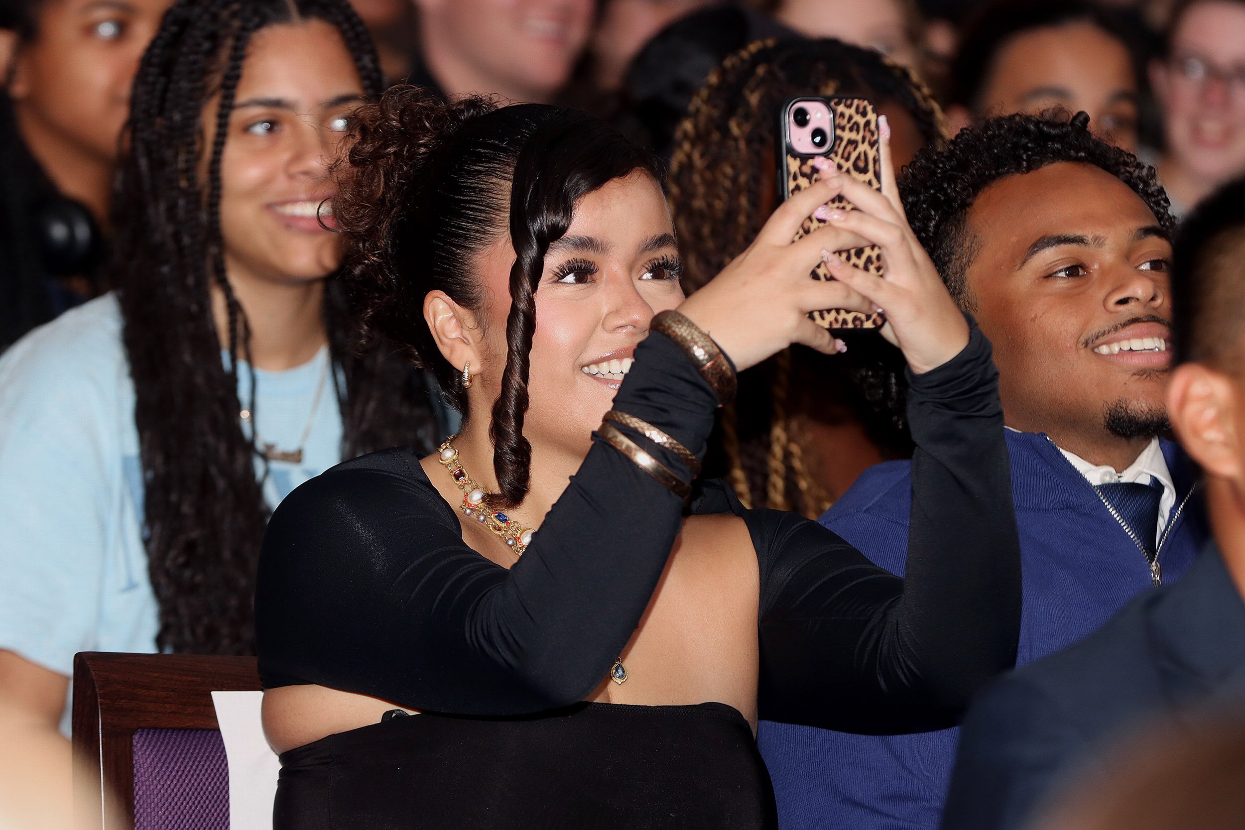 A young Latina woman with dark hair and dark eyes in a black dress is seated among a crowd indoors and takes a picture with her mobile phone of something off screen in front of her.