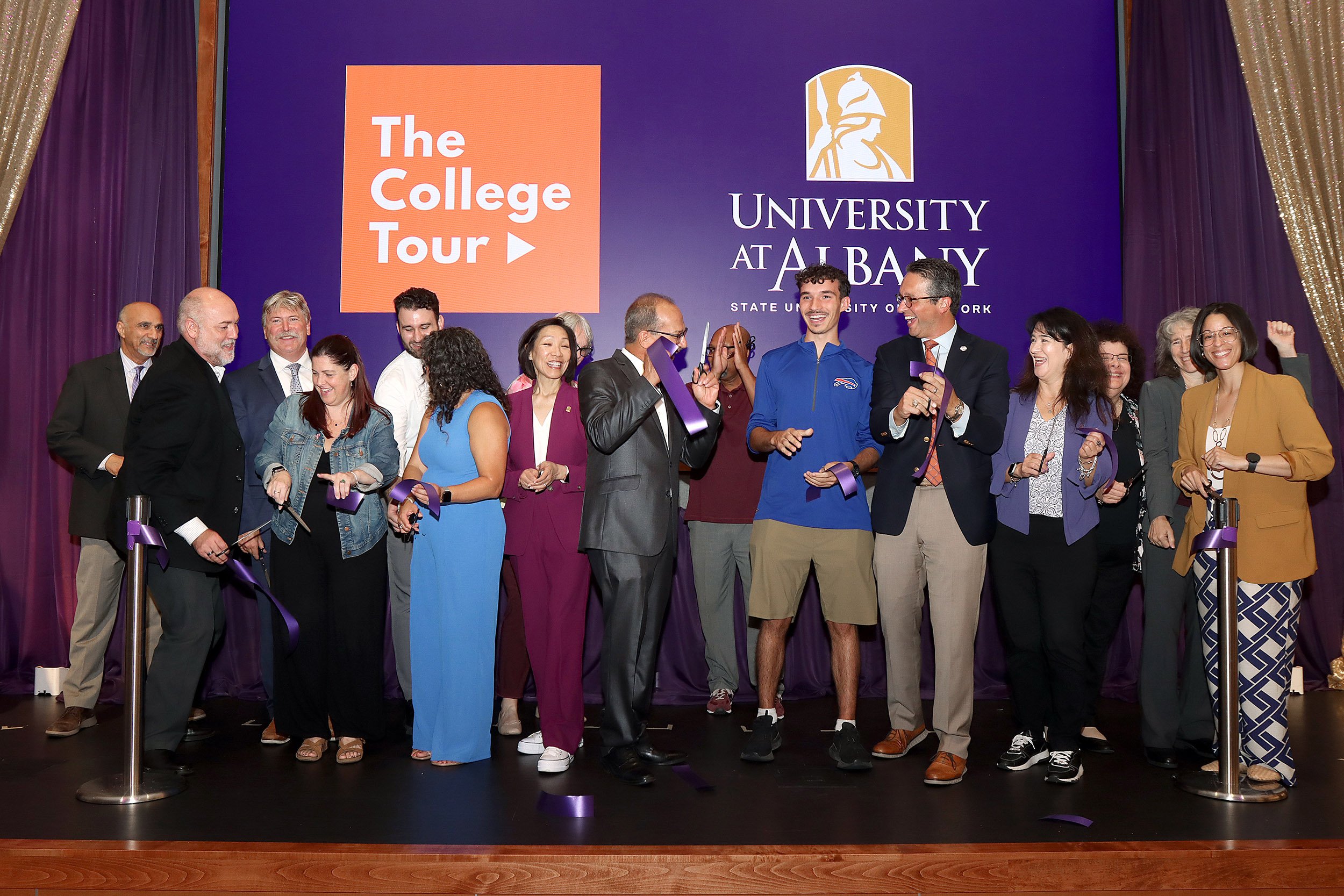 A group of people stand with scissors for a ribbon cutting that has just been completed indoors with the words "The College Tour" and "The University at Albany" on display behind them.