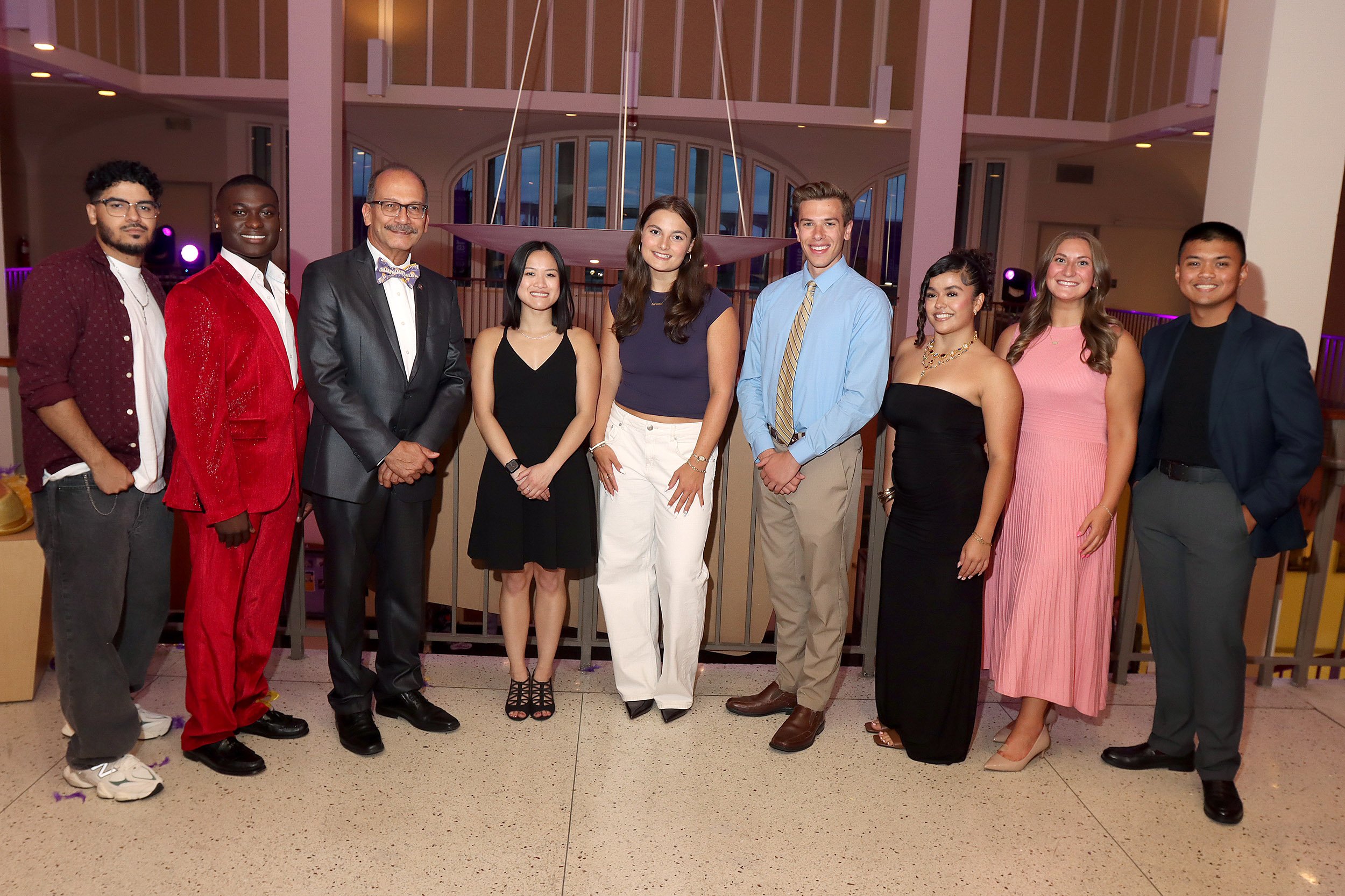 A crowd of students are joined by an older man in a bow tie at the top of a staircase in a posed picture indoors.