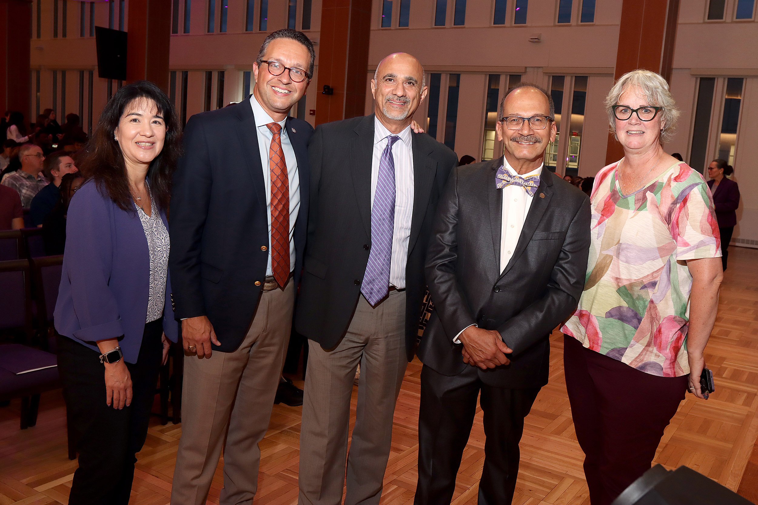 A group of two women and three men in formal business attire pose indoors for a photo inside of a ballroom with a wood floor and wood accents and tall windows in the background.
