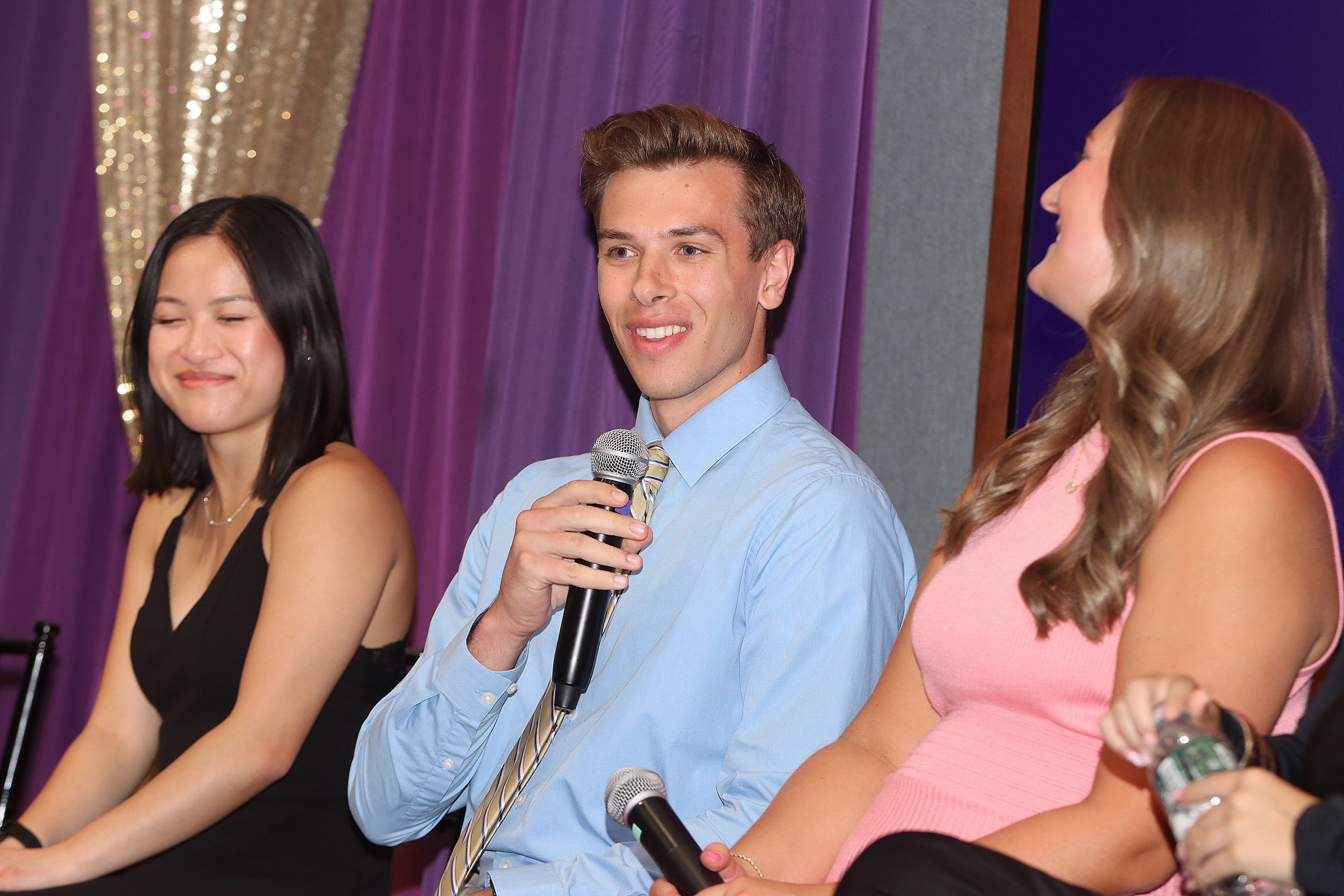 A young man with a blue shirt and tie holds a microphone while in between a young woman in a black dress with dark hair and a young woman in a pink dress with light hair indoors.