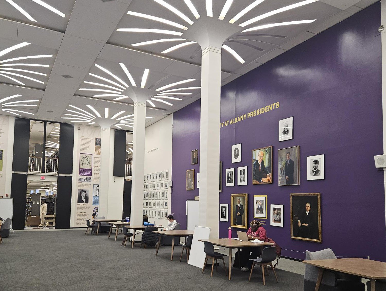 Students study at individual study tables in front of the a wall with paintings of UAlbany Presidents inside the University Library.