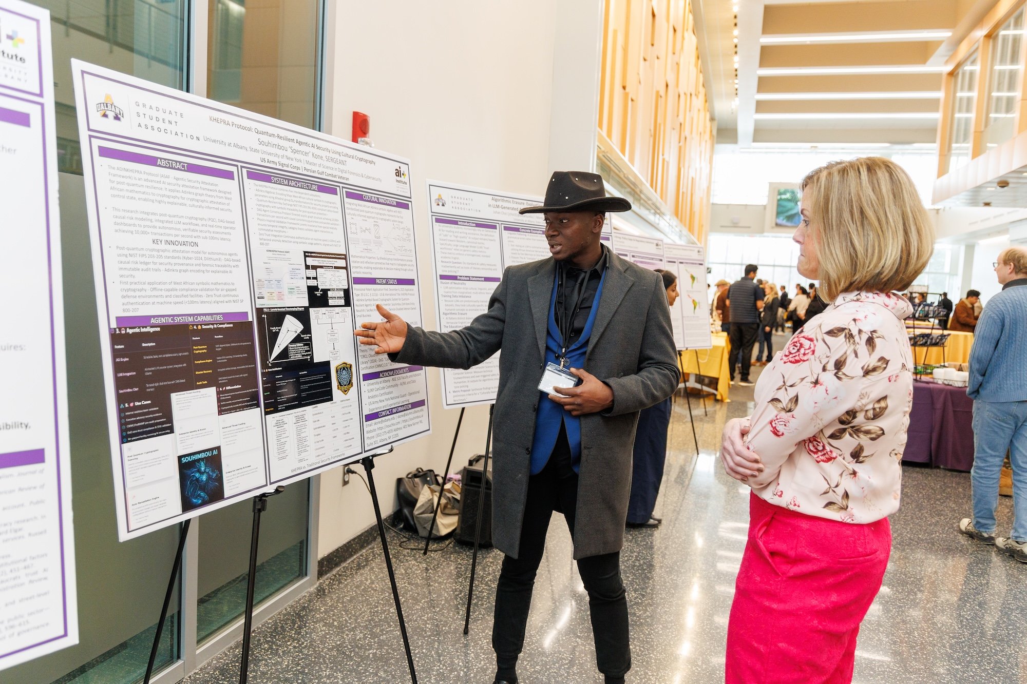 A man in a cowboy hat gestures toward a poster as he speaks to a woman watching nearby