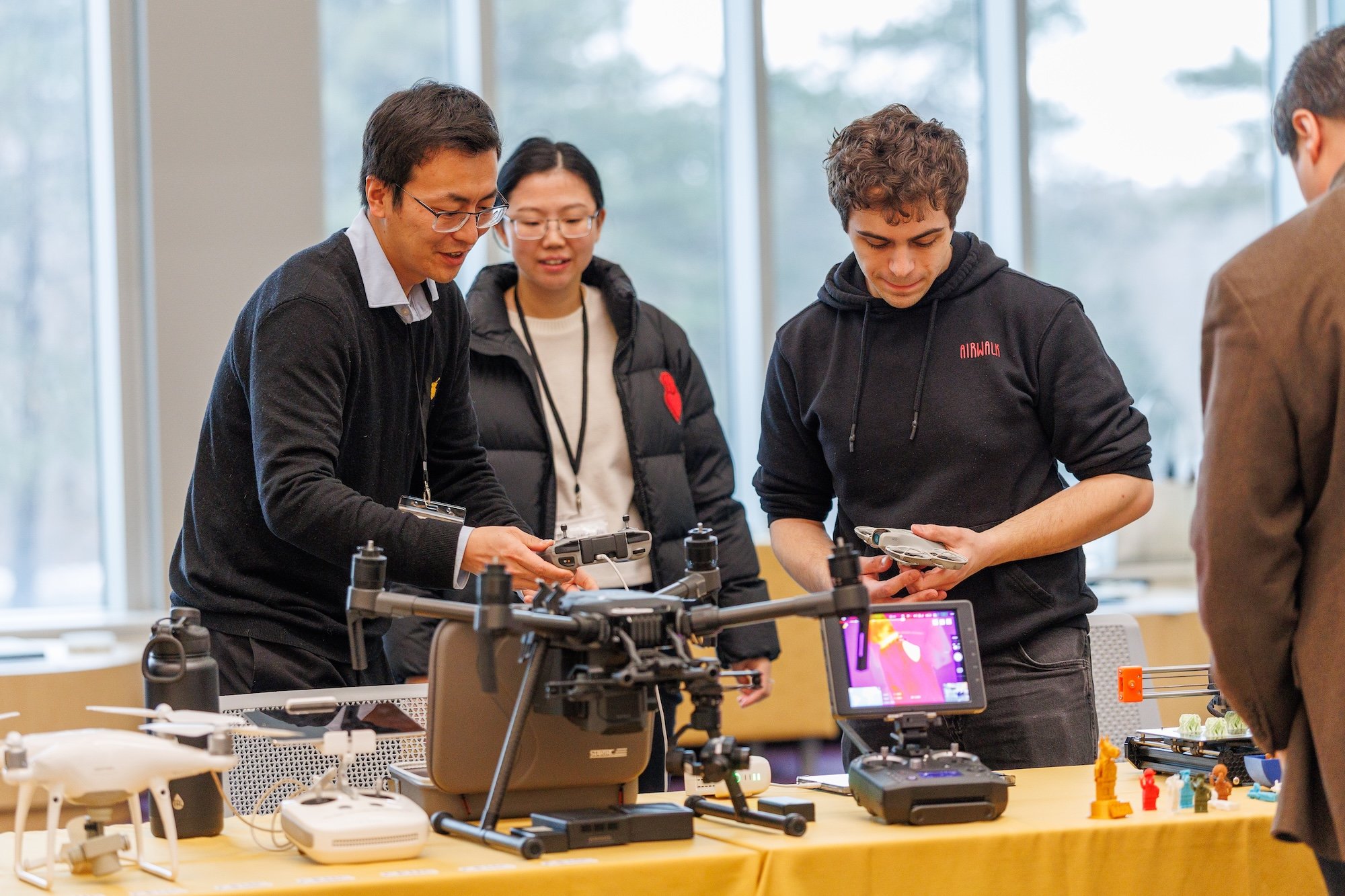 Two men gathered around a table topped with drones and other technology hold remote controllers as a woman looks on.
