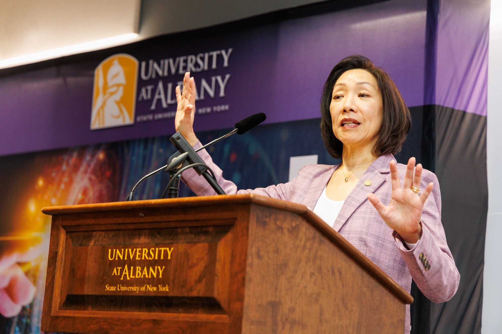 A woman in a lilac suit gestures in front of a wooden UAlbany podium in front of a purple and gold UAlbany logo