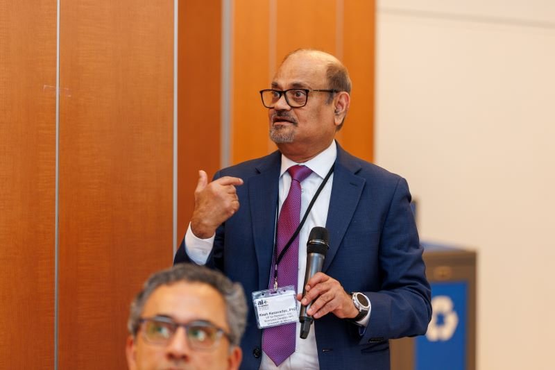 A man in a blue suit and purple tie gestures toward himself with one hand while speaking and holding a microphone in the other.