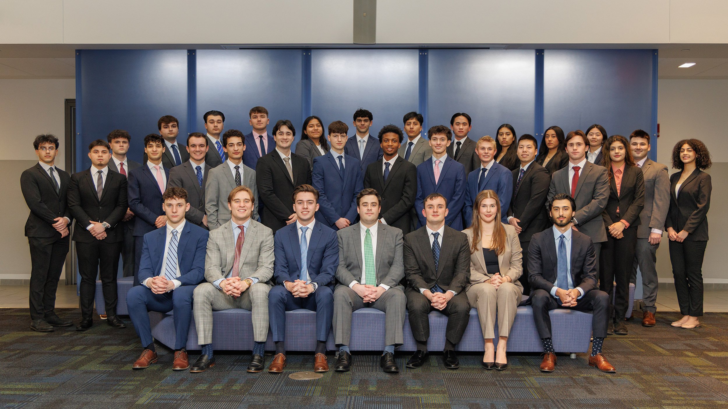 A large group of students are seated and standing wearing professional outfits in the Massry School of Business