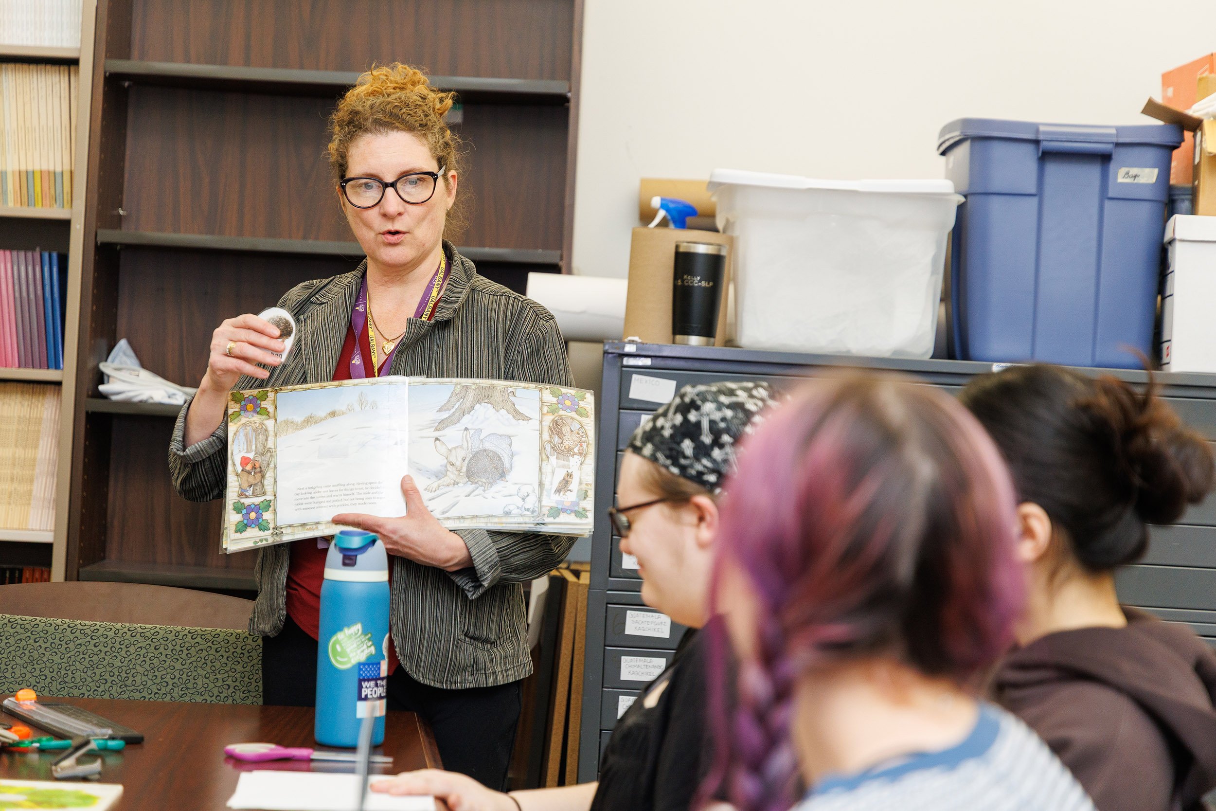 An instructor with red hair and glasses shows a book to a group of students in a conference room, demonstrating how to assemble adapted books.
