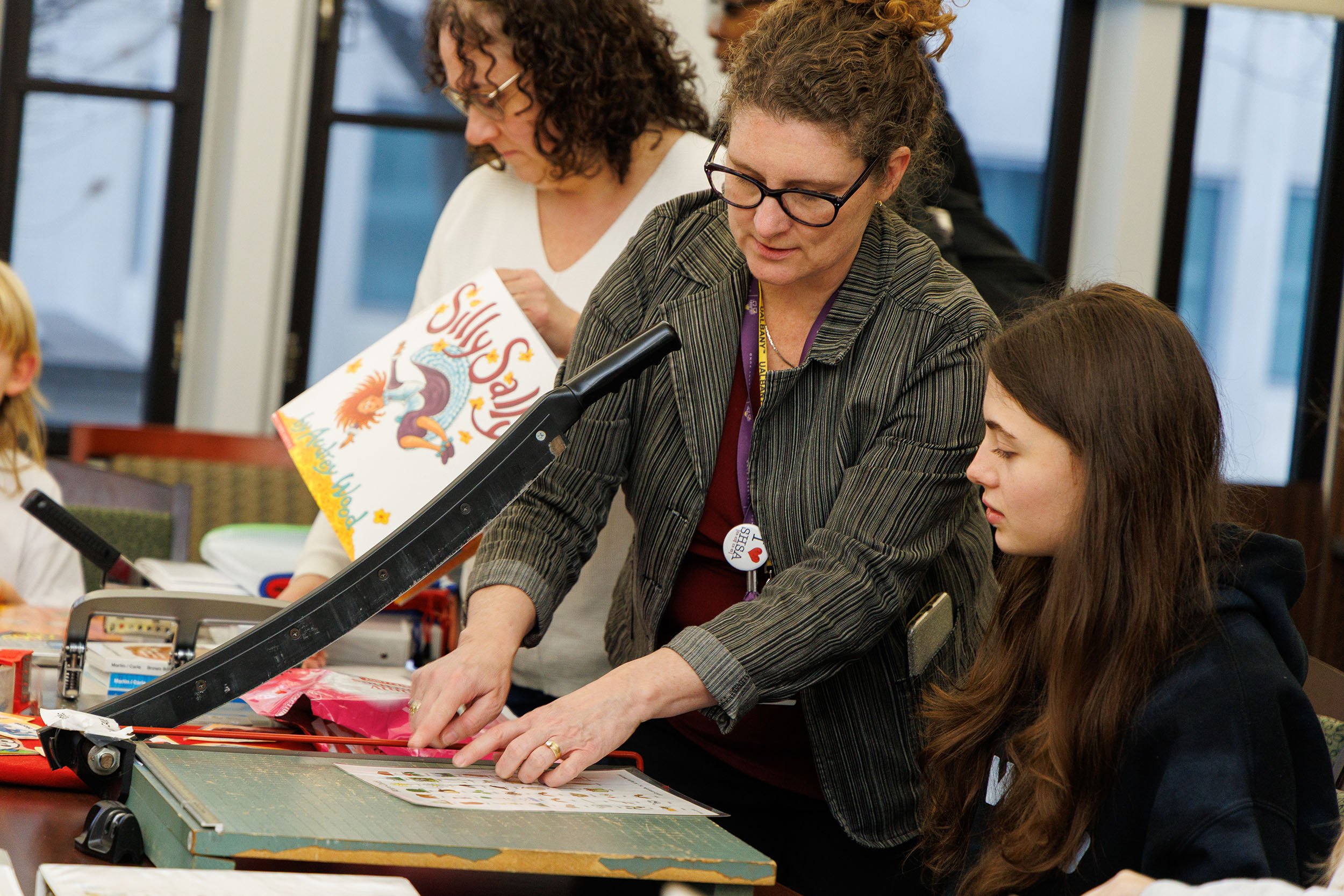 An instructor with red hair and glasses shows a book to a group of students in a conference room, demonstrating how to assemble adapted books.