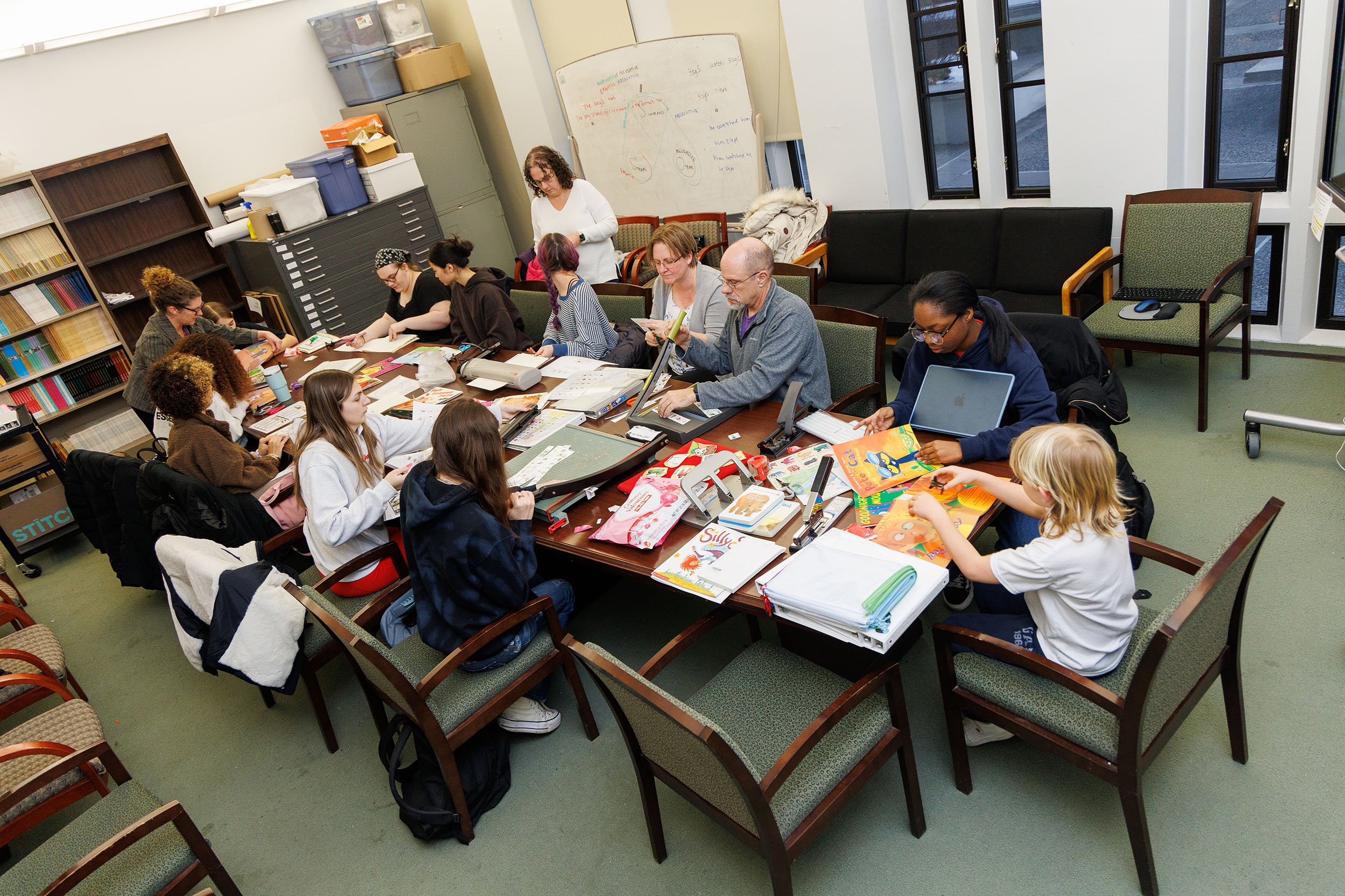 A group of students, instructors and volunteers work together at a long table assembling adapted children’s books.