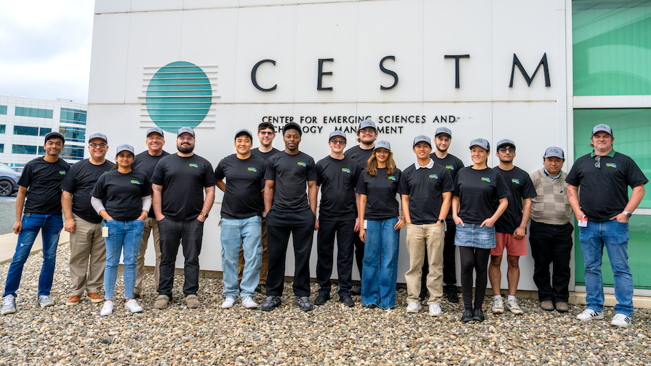 More than a dozen people gather for a group photo outside of a building with a sign that says CESTM Center for Emerging Sciences and Technology Management