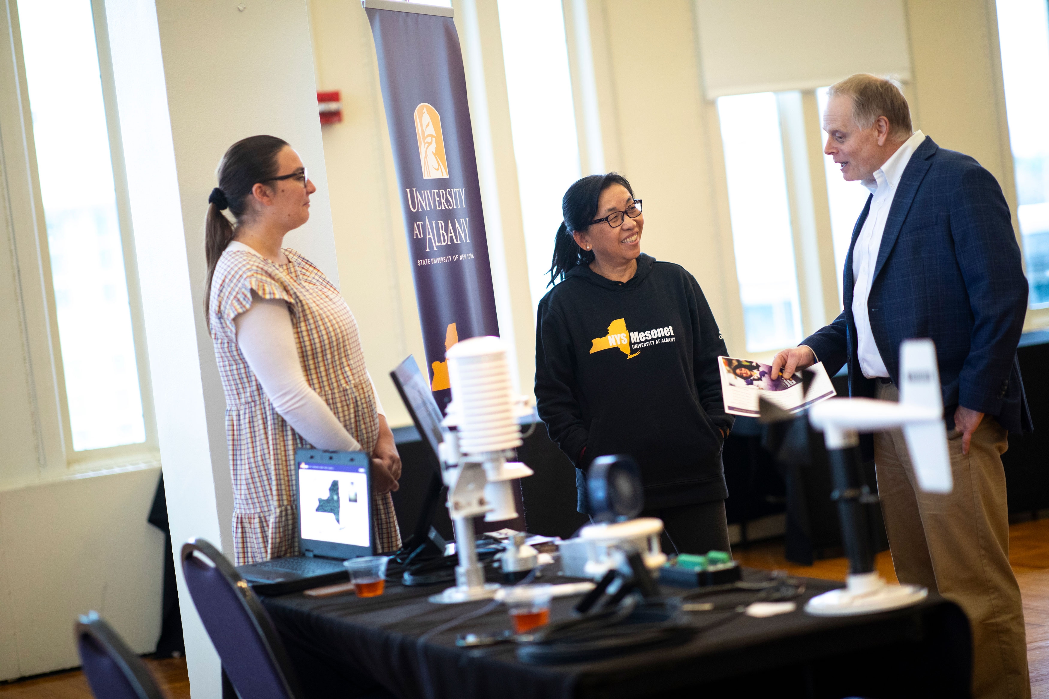 Two New York State Mesonet researchers staff a table at R&E Week 2023. An attendee is speaking with them and looking over the technology demonstration they have set up.