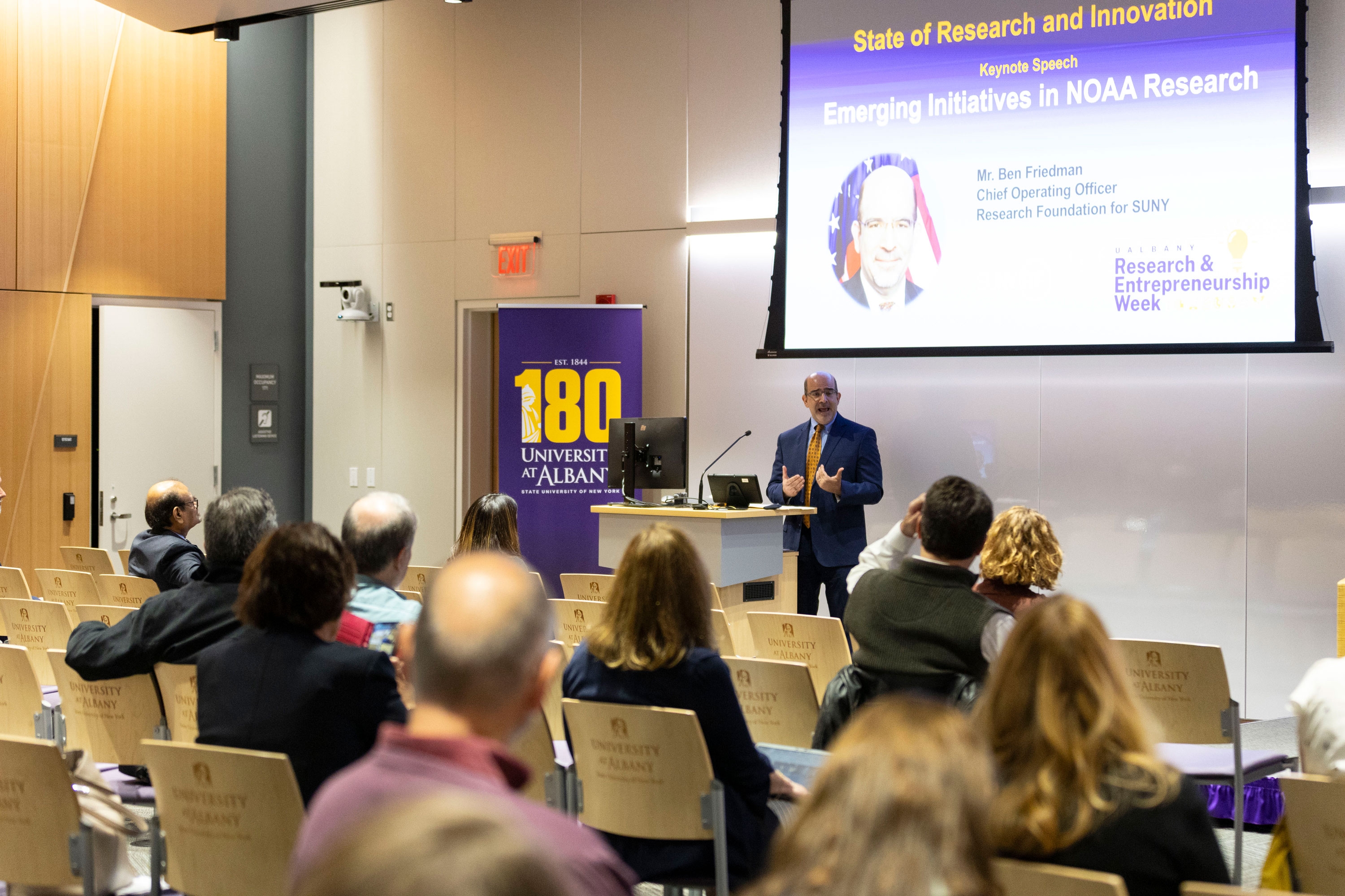 Benjamin Friedman, Chief Operating Officer of the Research Foundation for SUNY, speaks in front of a seated crowd at R&E Week 2024. The slide projected at a screen at the front of the room includes the title, "State of Research and Innovation, Keynote Speech, Emerging Initiatives in NOAA Research."