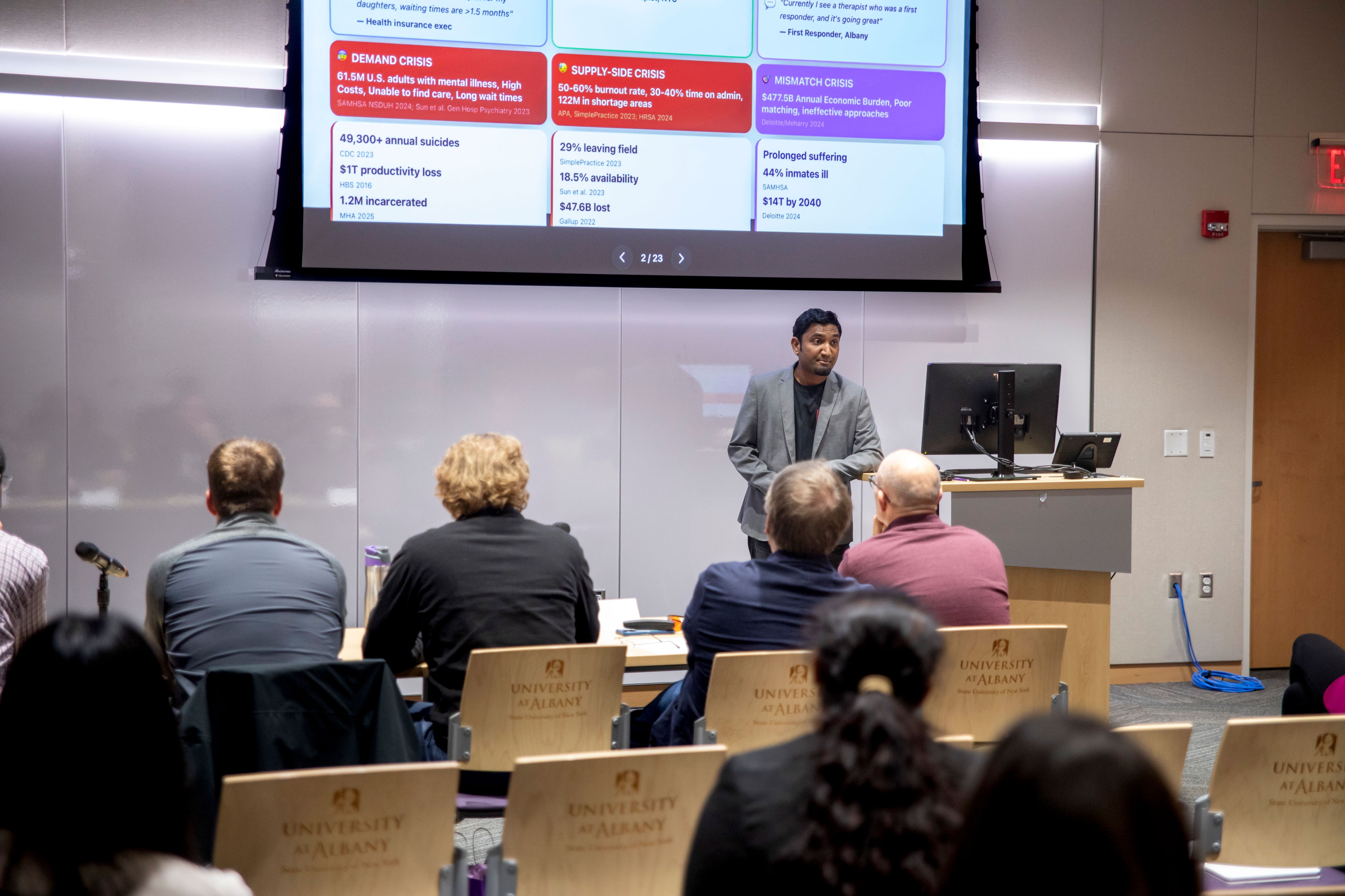A presenter leans on the podium while speaking to a seated audience at R&E Week 2025. The slide displaying via projector includes data on the crises his product is built to solve.