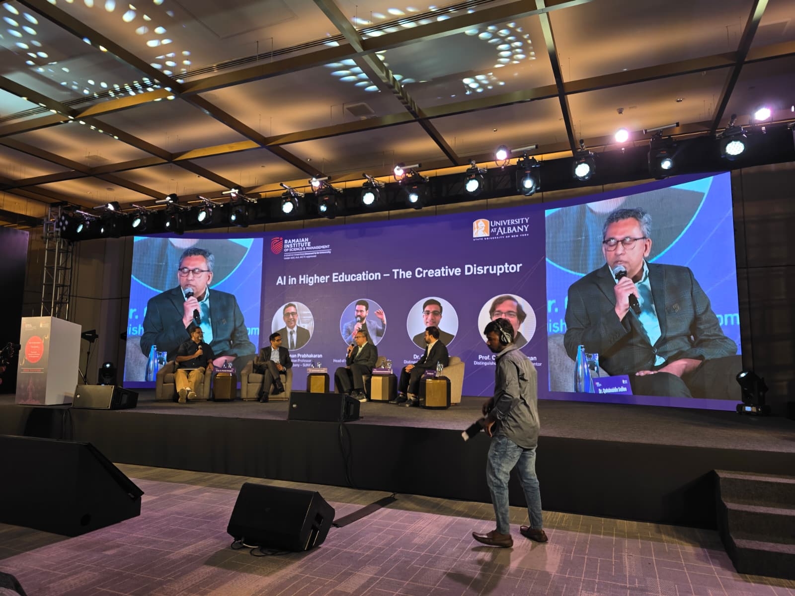 Four officials from UAlbany and RISM sit on stage and engage in a panel discussion. Behind them, a screen shows their names, titles and photos, as well as the words, "AI in Higher Education - the Creative Disruptor."