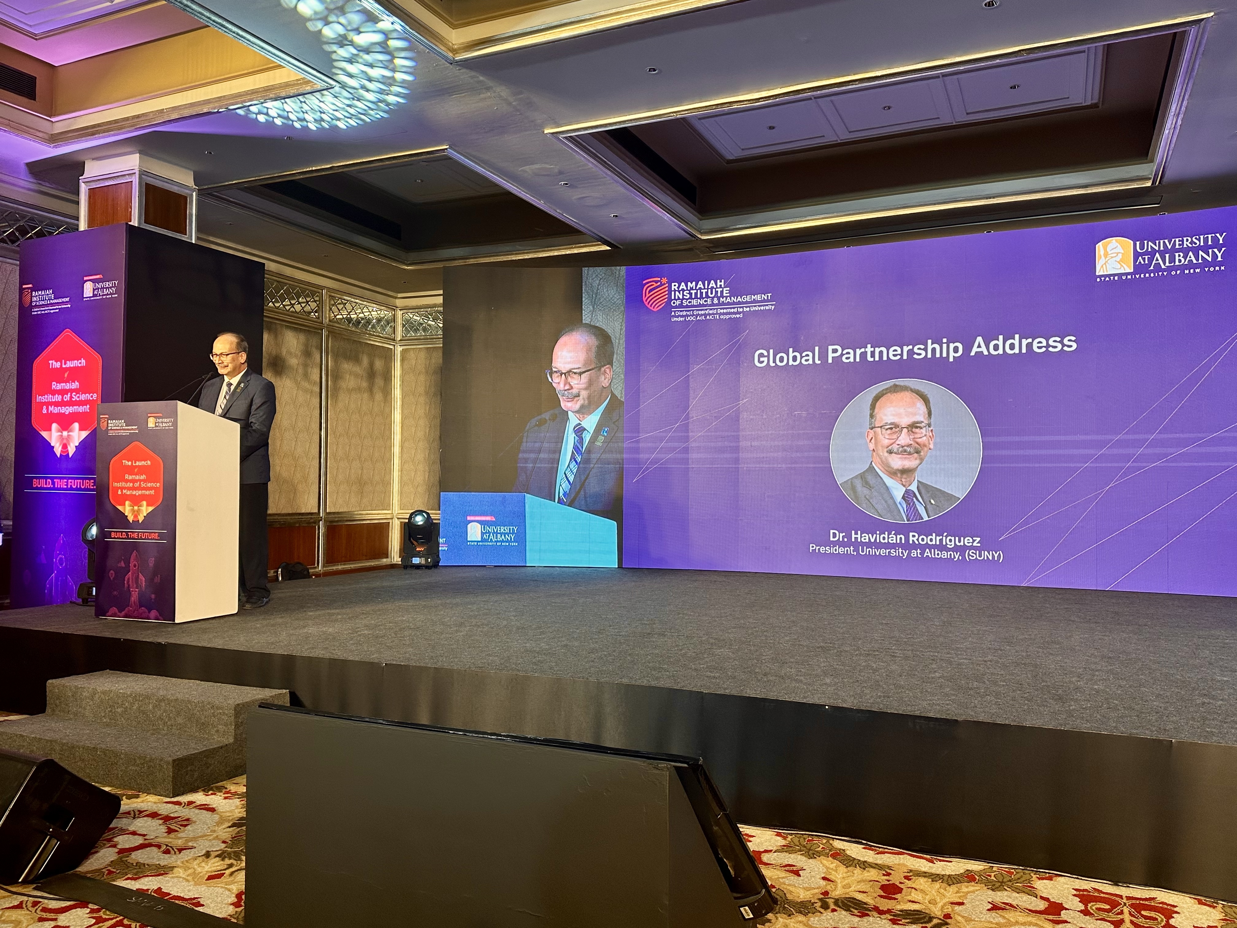 UAlbany President Dr. Havidán Rodríguez speaks at a podium on stage. Behind him, a screen shows his name, title and photo, as well as the words, "Global Partnership Address."