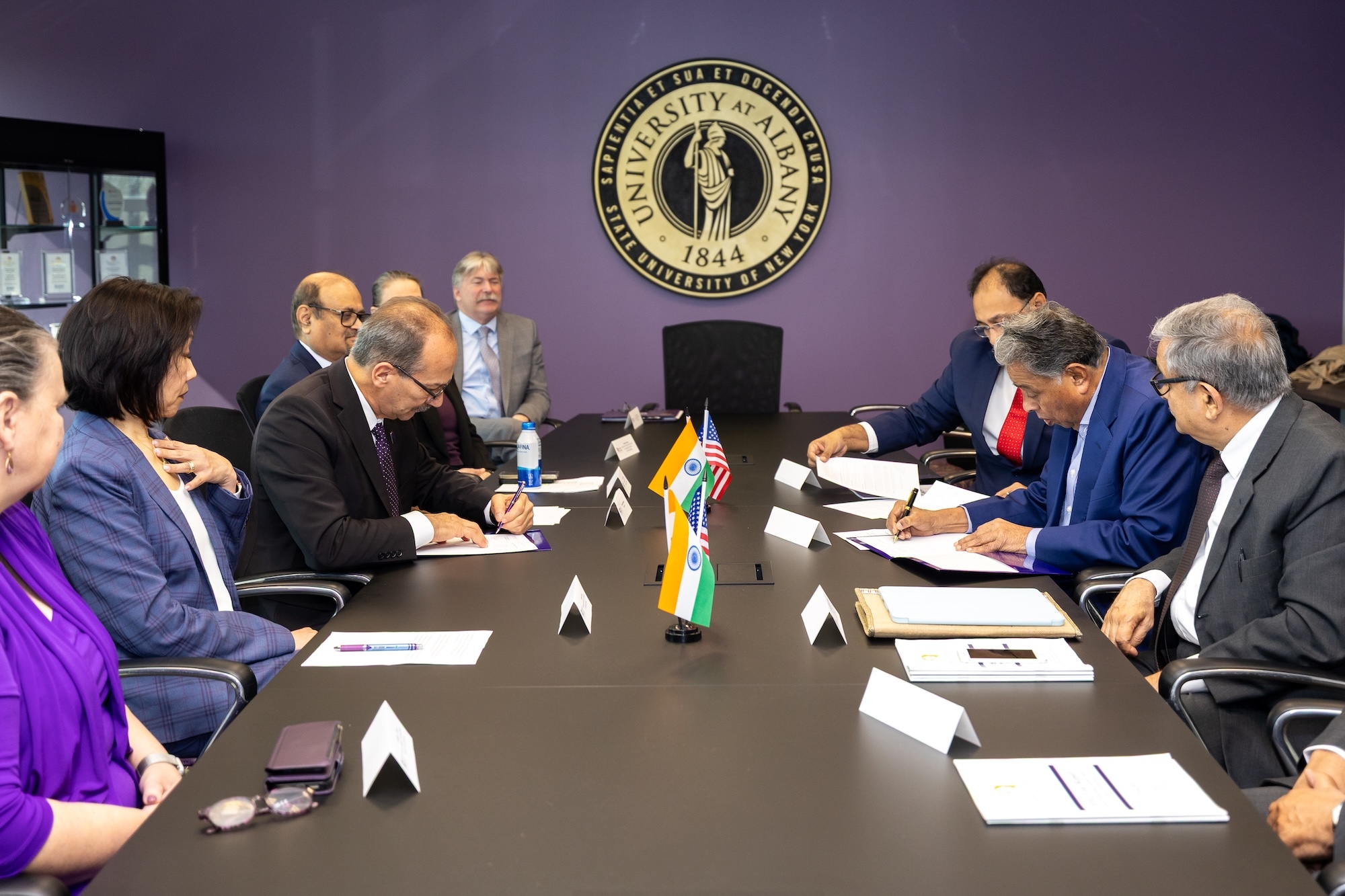 Leaders from UAlbany and RISM gather around a conference table to sign their collaboration agreement. The UAlbany seal is mounted on the wall behind them and the American and Indian national flags are positioned on the table.