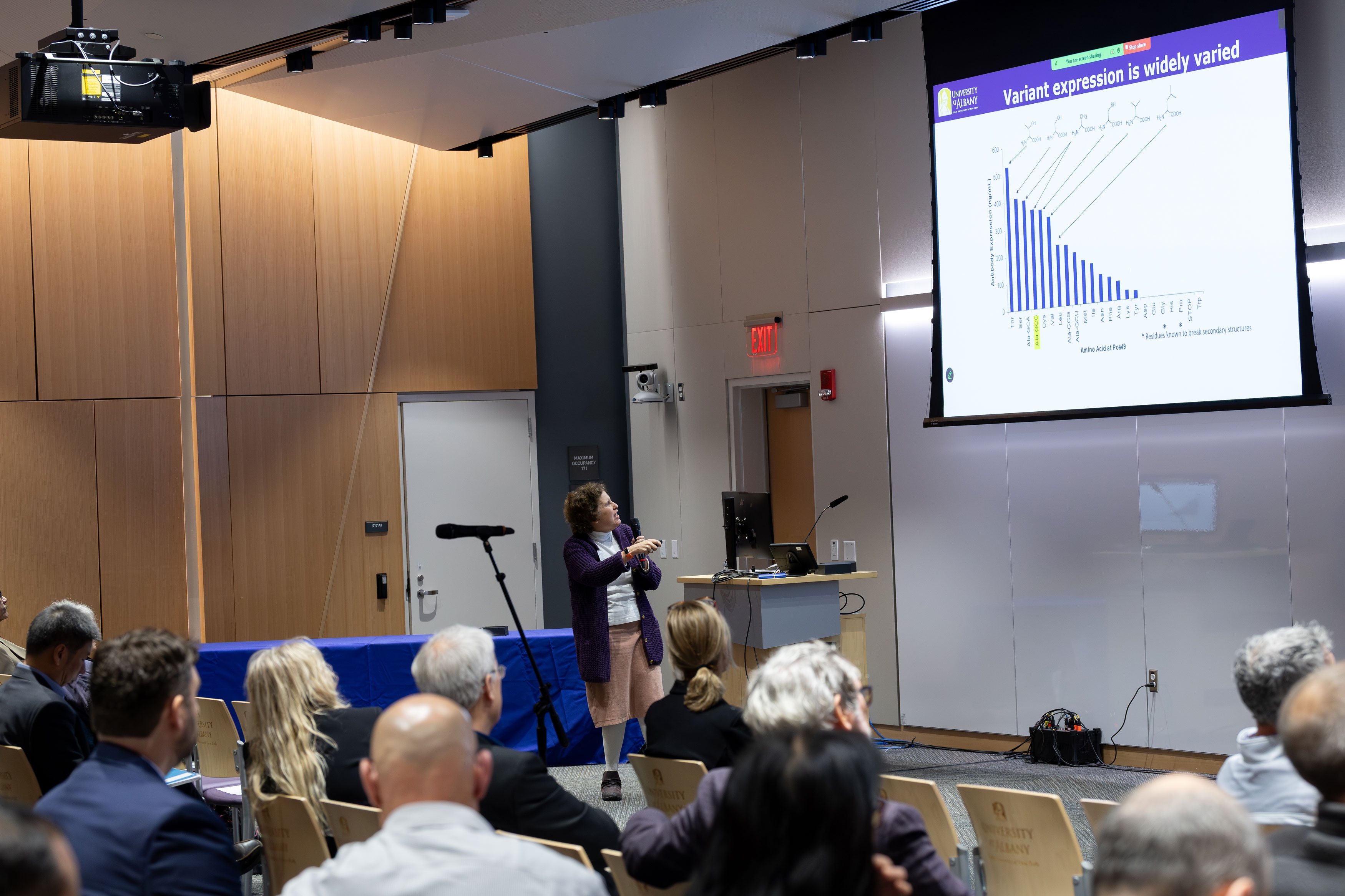 A presenter uses a handheld clicker to change slides while speaking into a microphone in front of an audience at R&E Week 2025. The slide displaying includes the title, "Variant expression is widely varied," and a bar chart.