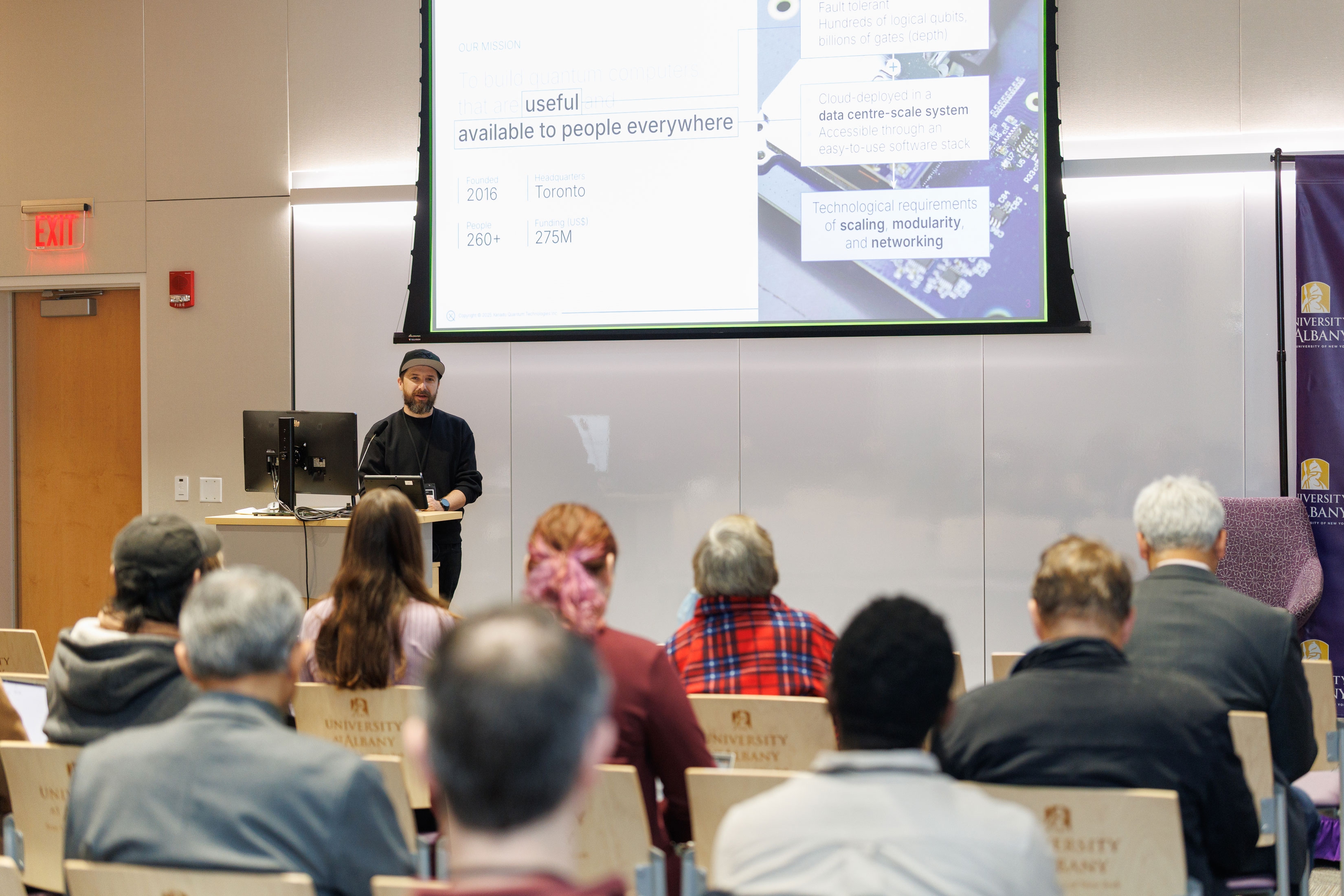 A presenter speaks in front of a seated crowd at R&E Week 2025. The slide displaying on the projector screen includes data points and quotes.