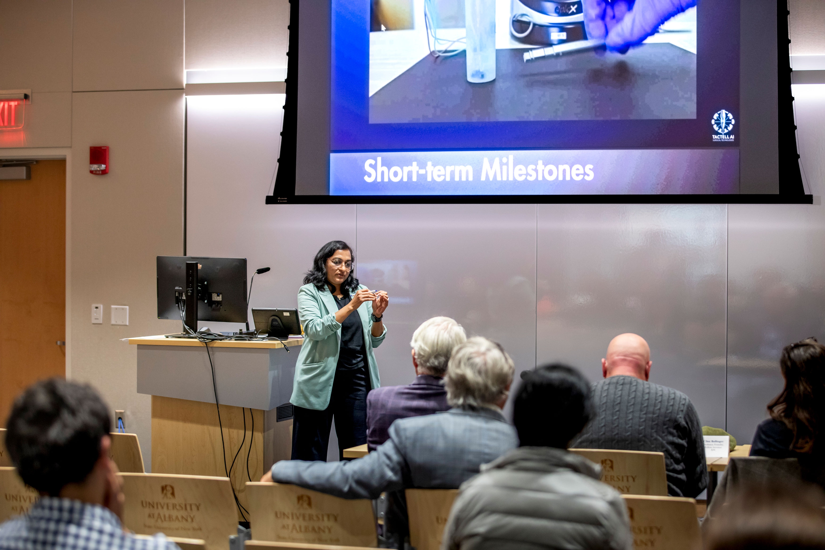 A presenter gestures while speaking to a seated audience at R&E Week 2025. The slide displayed via projector includes the title "Short-term Milestones."