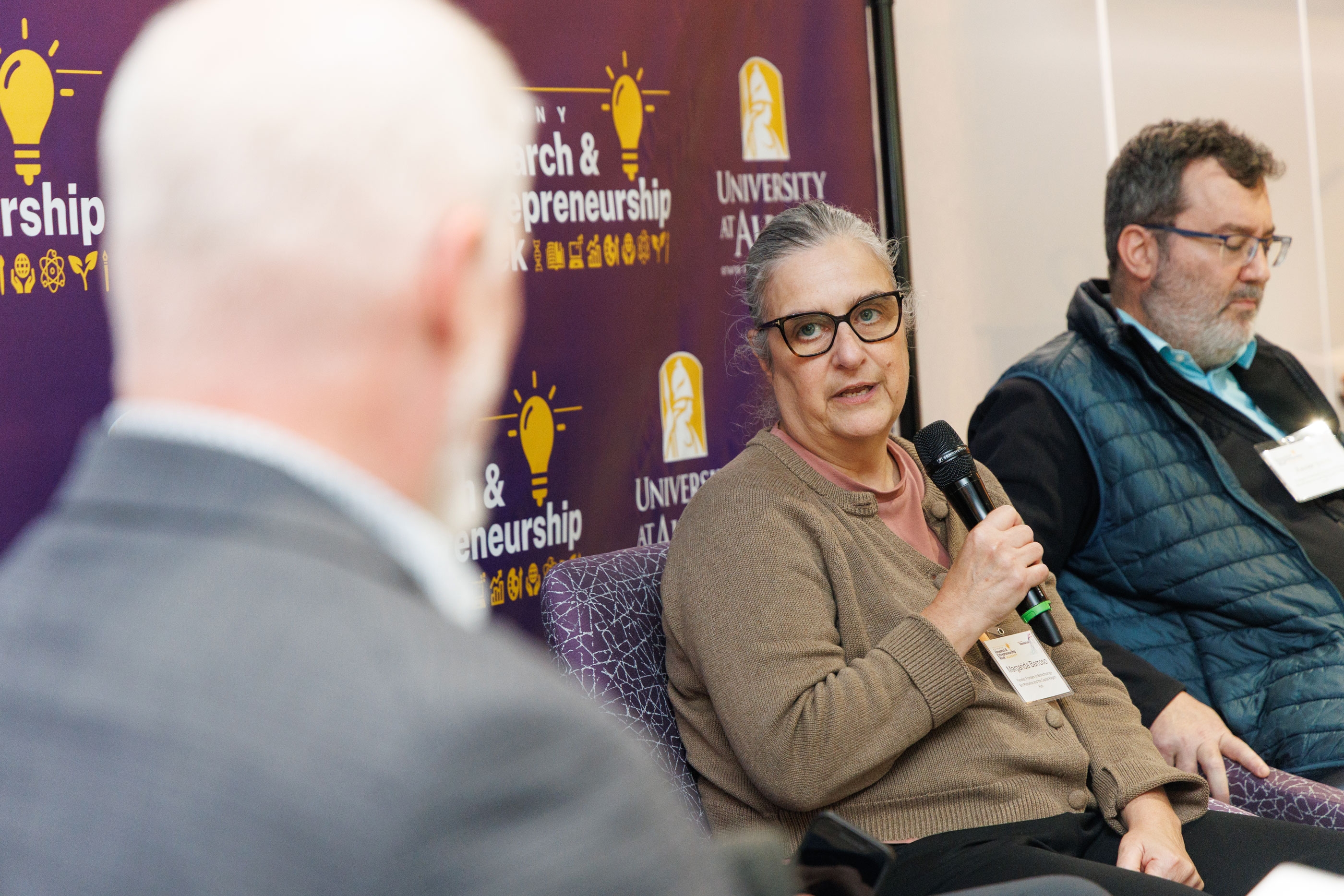 A panelist speaks into a handheld microphone while looking at the moderator. A second panelist sits beside her.