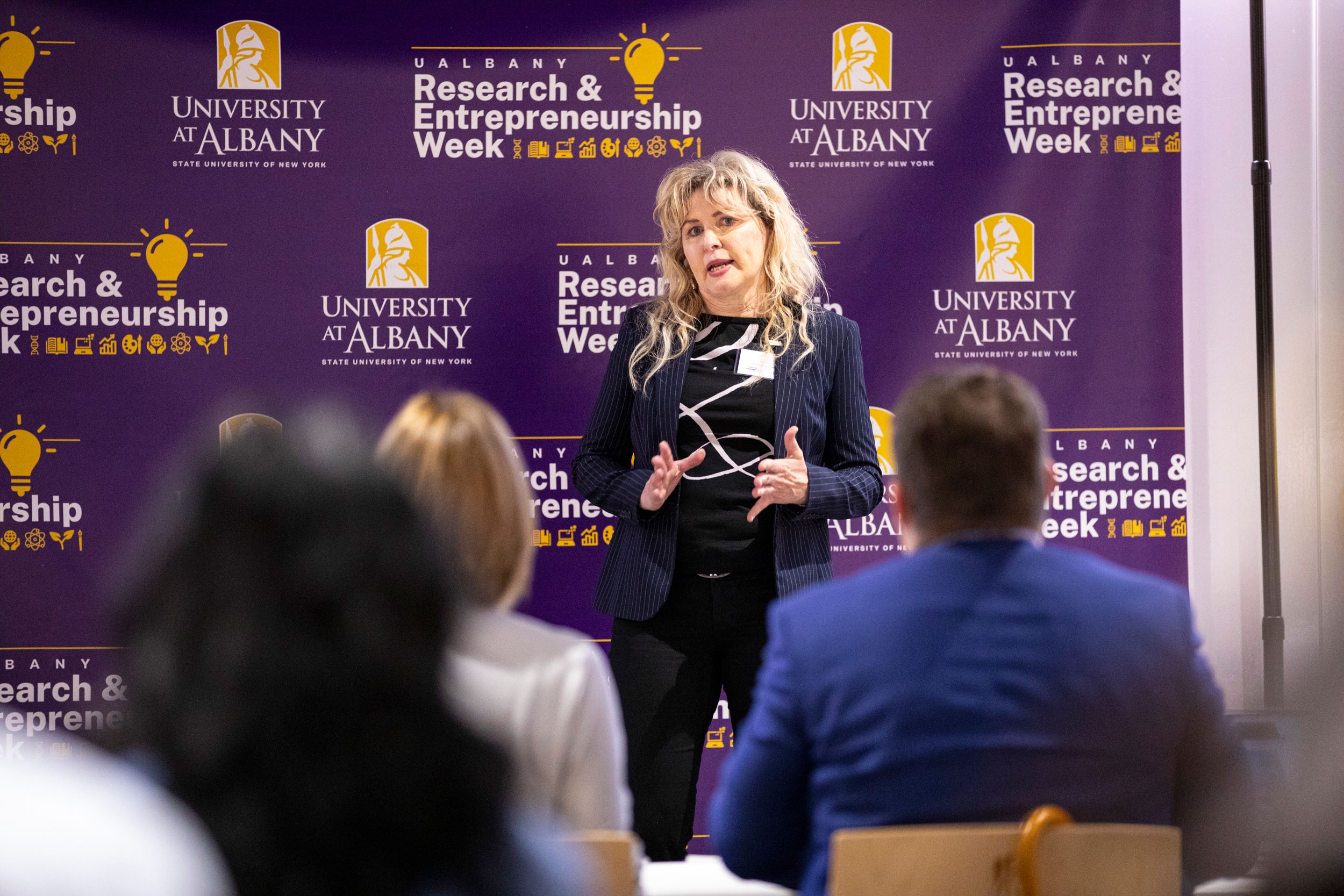 A researcher stands in front of an R&E Week banner and pitches her invention to a panel of judges.