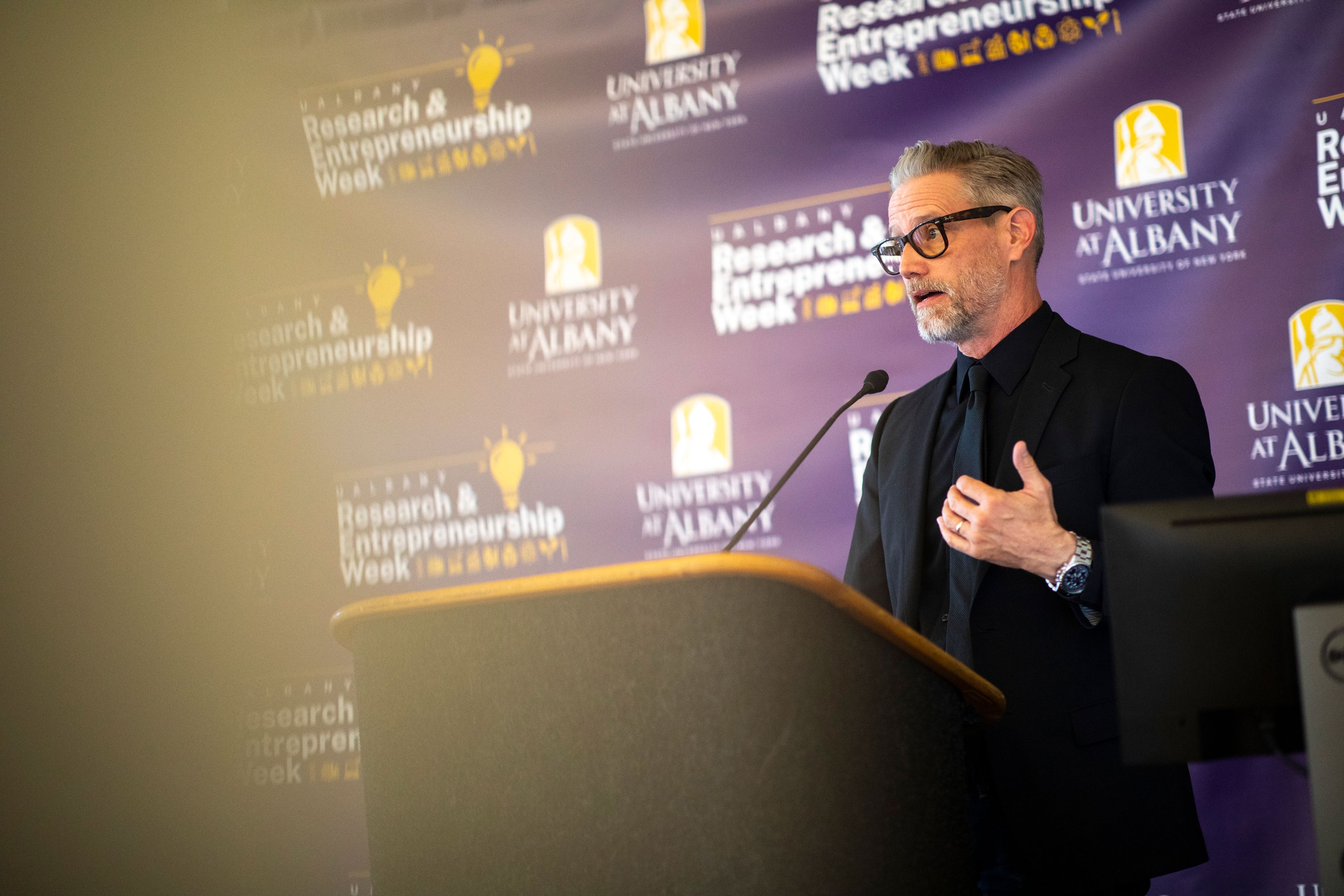 A researcher stands at a podium and speaks during R&E Week 2023. He stands in front of a purple backdrop with the event logo.