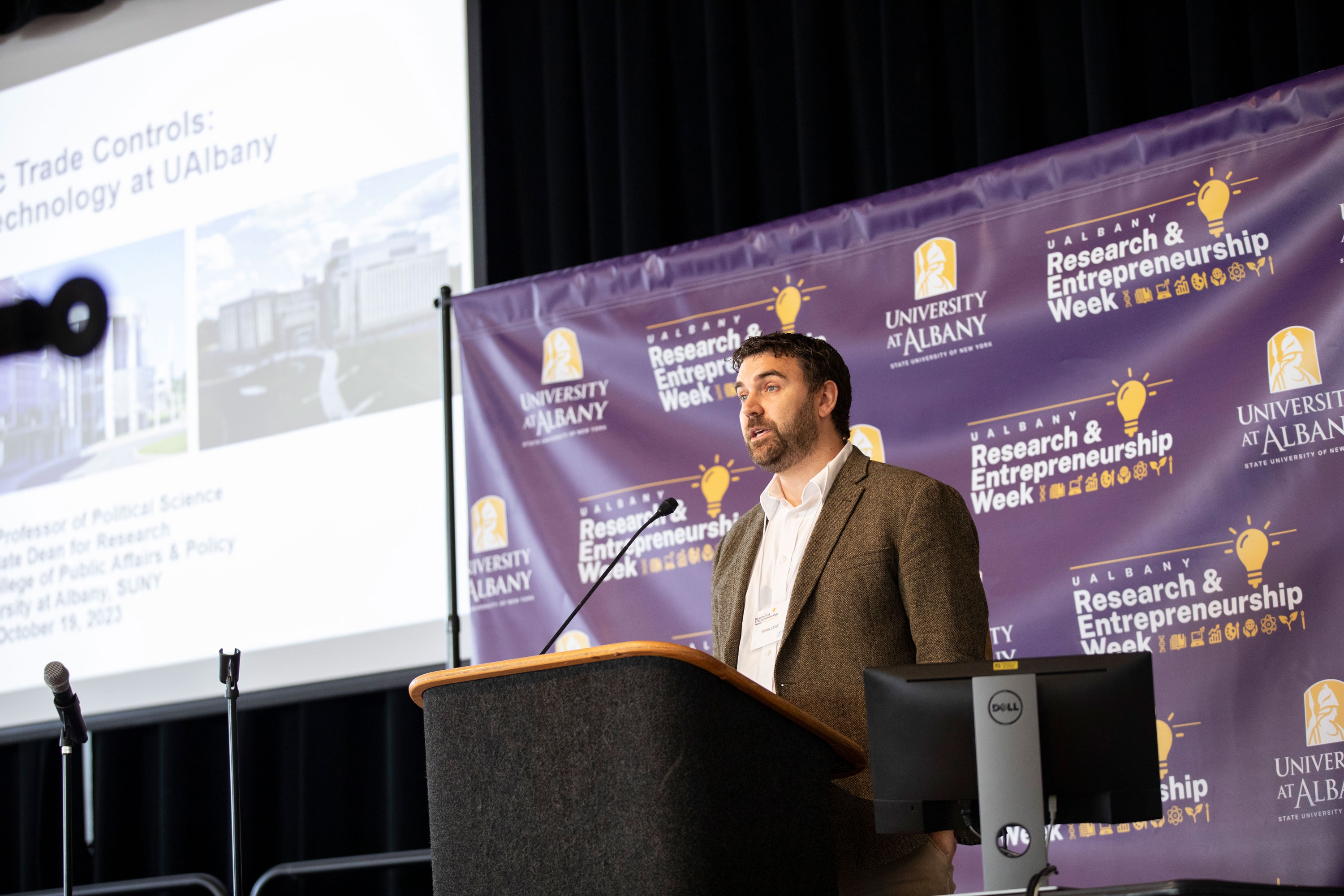 A researcher speaks from a podium in front of an R&E Week 2023 backdrop. The slideshow being projected on the screen mentions trade controls.