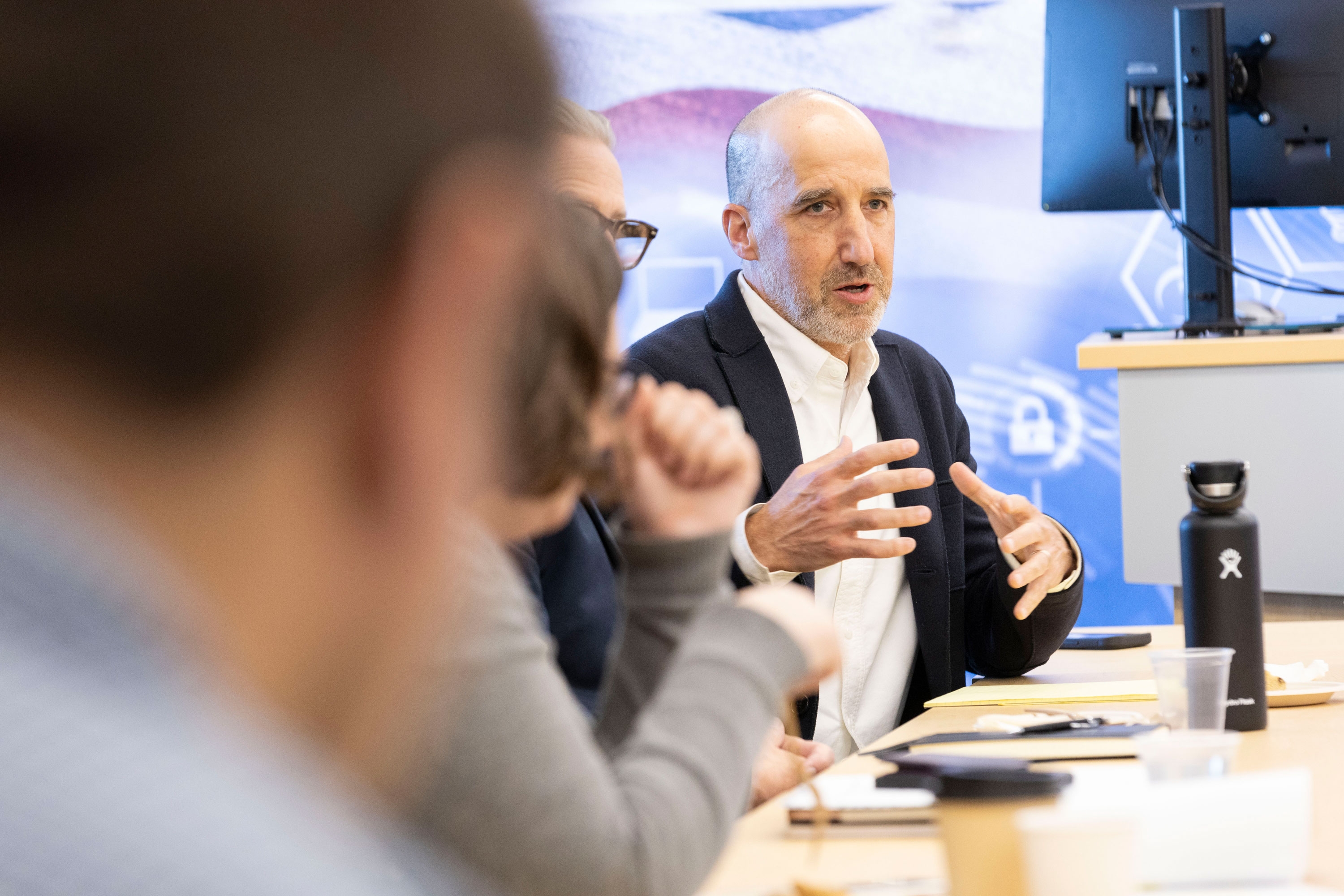 A researcher sitting at a table with other R&E Week 2024 attendees gestures with his hands while speaking.