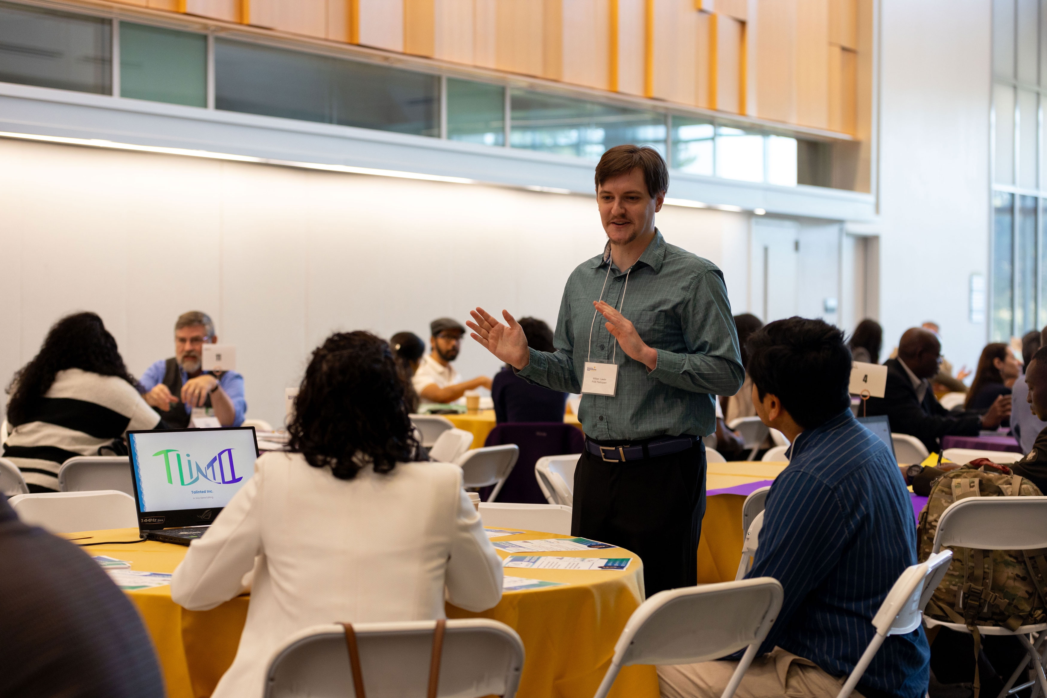 A researcher stands, smiles and gestures while speaking to two seated students during a RISE pitch session.