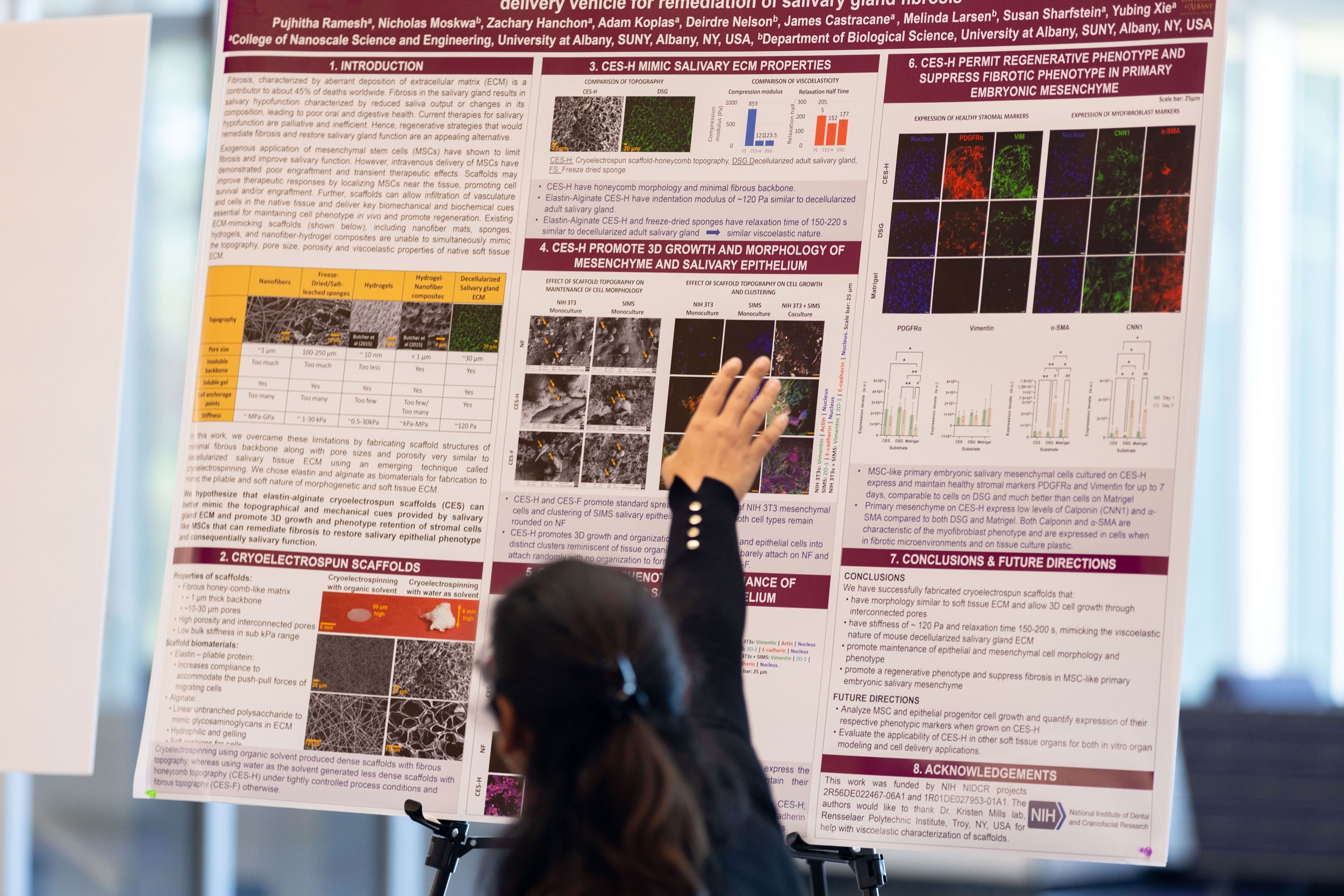 A researcher sits at a table and gestures behind her towards a research poster at R&E Week 2024.