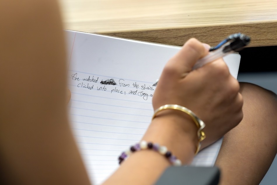 A girl's hand holds a pen over a notebook containing handwritten sentences.